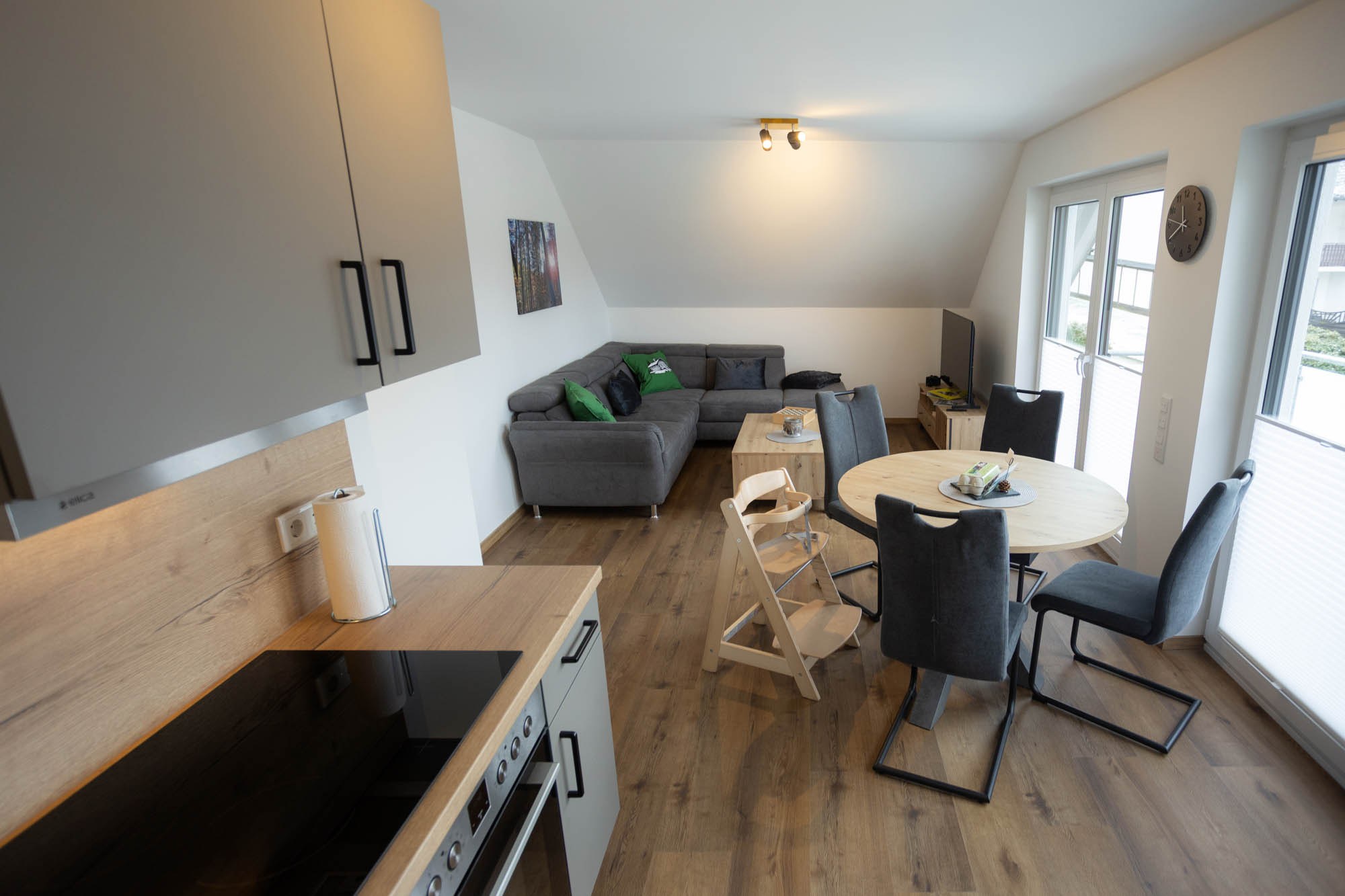 Moderner Wohn- und Essbereich mit graßer Eckcouch, Holz-Esstisch mit vier Stühlen, Kinderhochstuhl, und Küchenzeile mit Herd und Ofen.