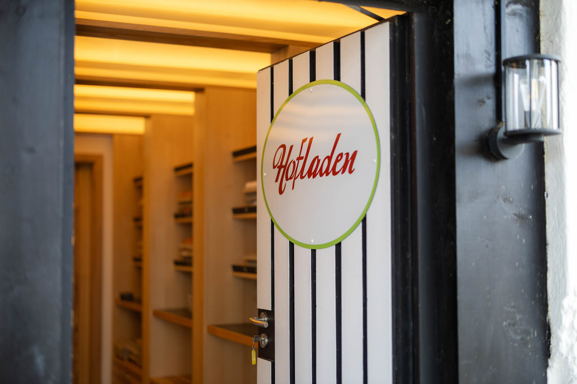 Offene Tür mit Schild „Hofladen“ zeigt Innenraum mit Regalen und warmem Licht.