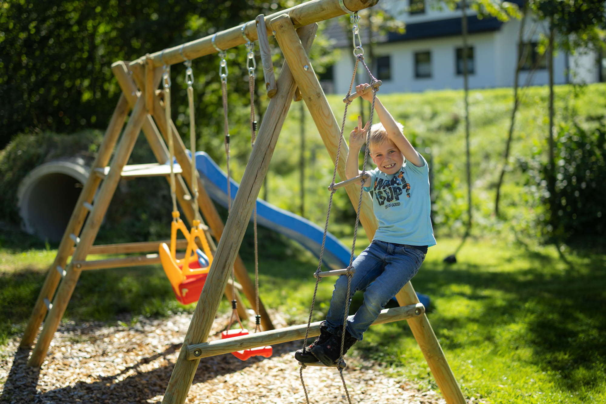 Ein Junge in einem hellblauen T-Shirt und Jeans spielt auf einer Seilleiter an einem Holzspielgerät im sonnigen Garten.