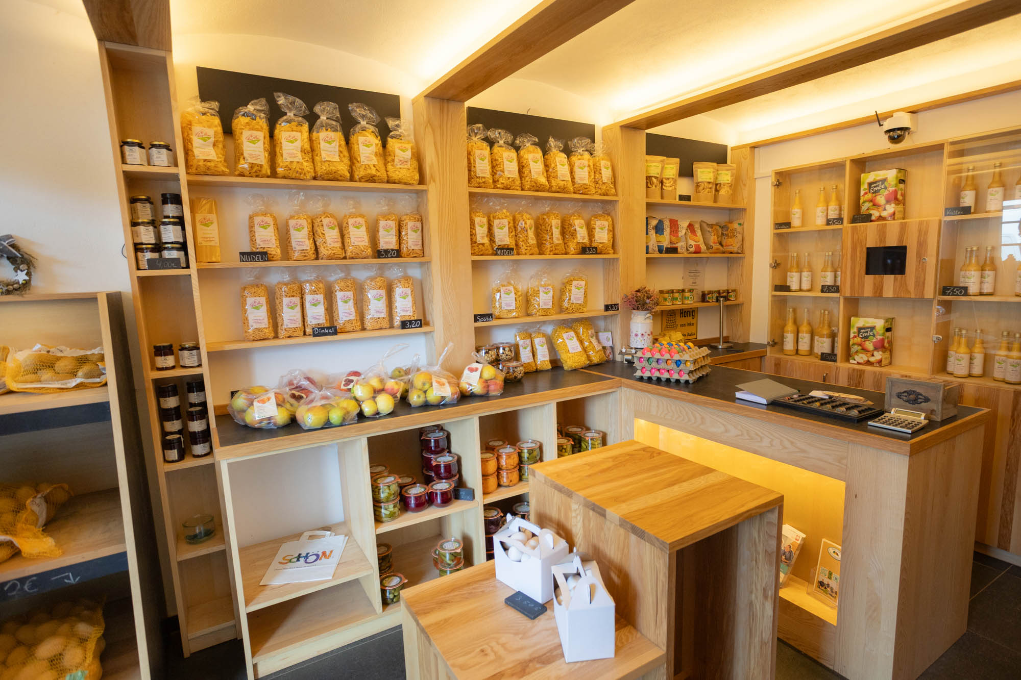 Kleiner Hofladen mit Holzregalen, gefüllt mit Nudeln, Obst, Marmeladen, Eiern und Getränken.