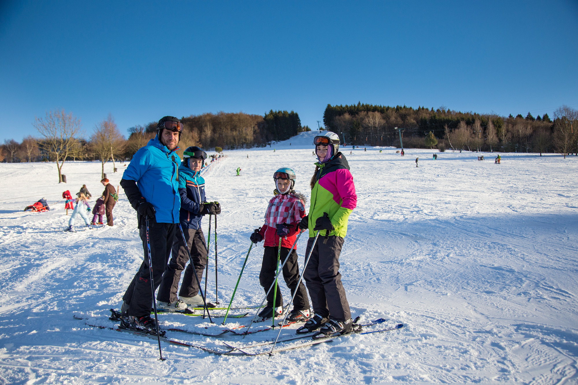 Vier Skifahrer in bunter Winterkleidung posieren auf einer verschneiten Piste mit blauem Himmel und Wald im Hintergrund.
