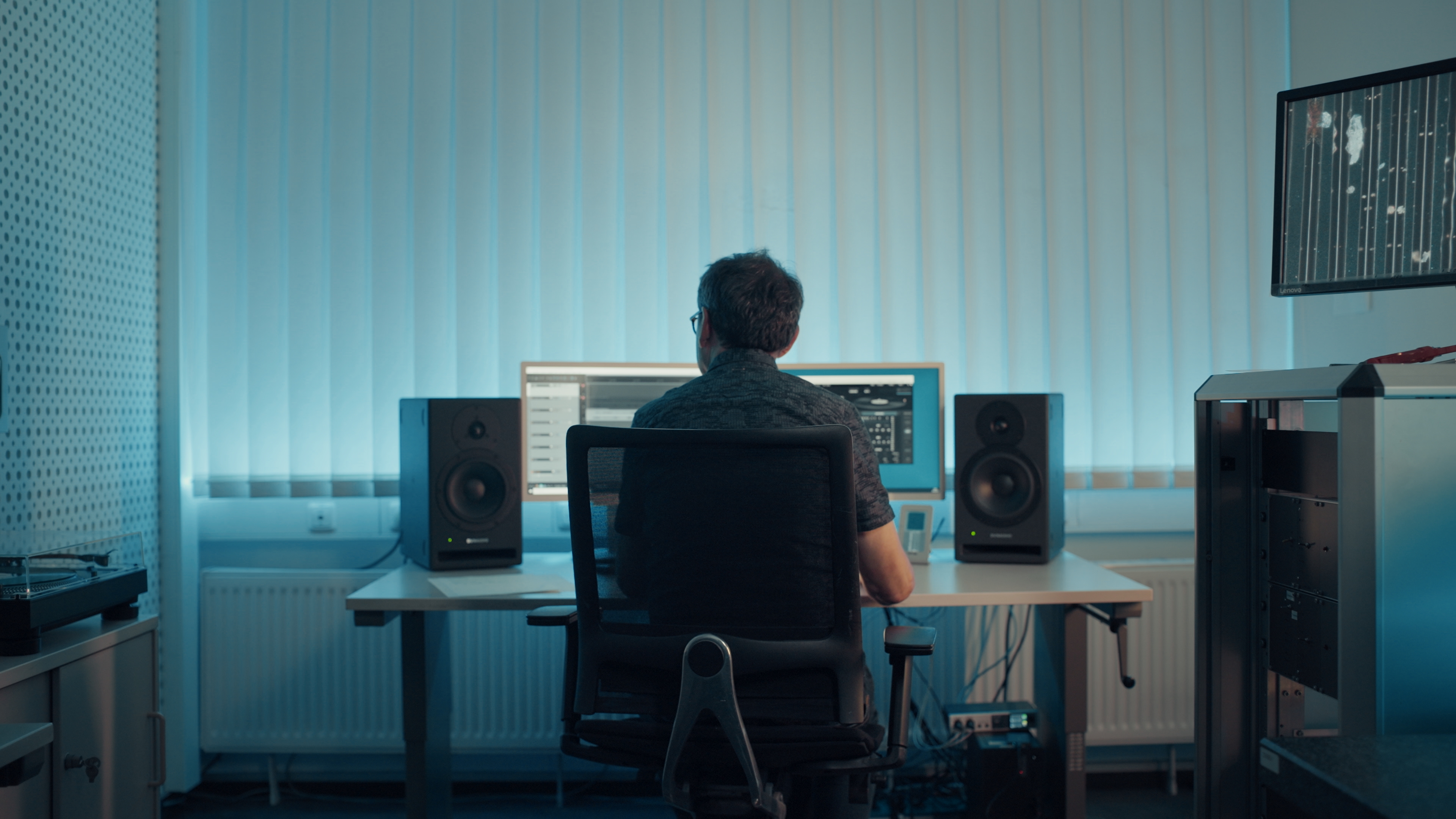 A man sits in a black mesh office chair and works on a computer with two monitors, flanked by speakers on either side, in a dimly lit room.
