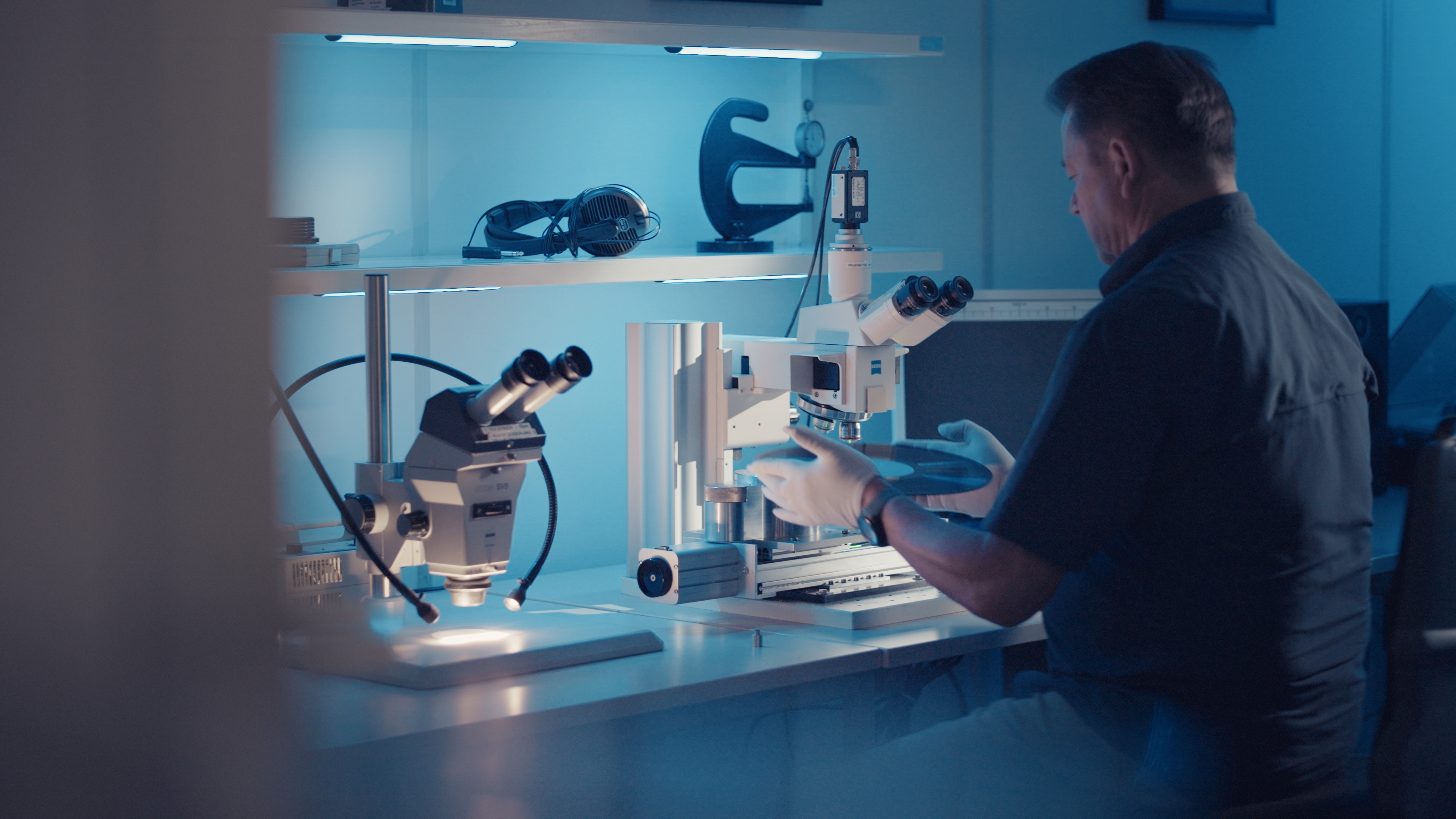 A man wearing gloves is handling a round object under a microscope in a blue-lit laboratory.