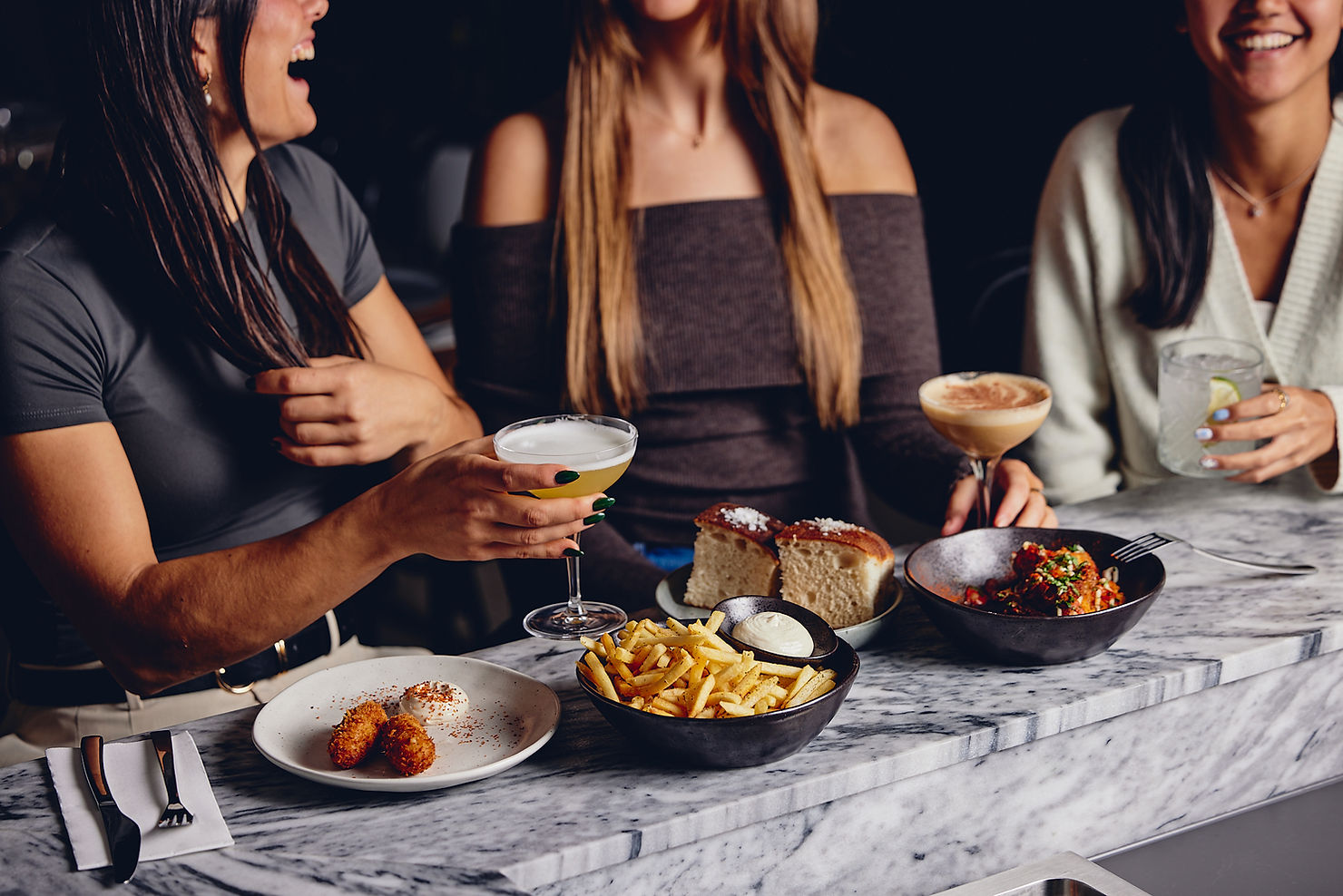 Three females enjoying cocktails and snacks at a marble bar.