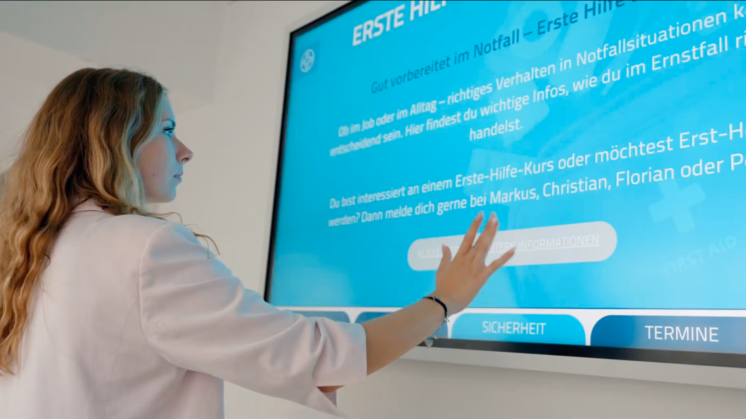 Frau mit langen lockigen Haaren bedient ein großes Touchscreen-Display mit Informationen zur Ersten Hilfe.