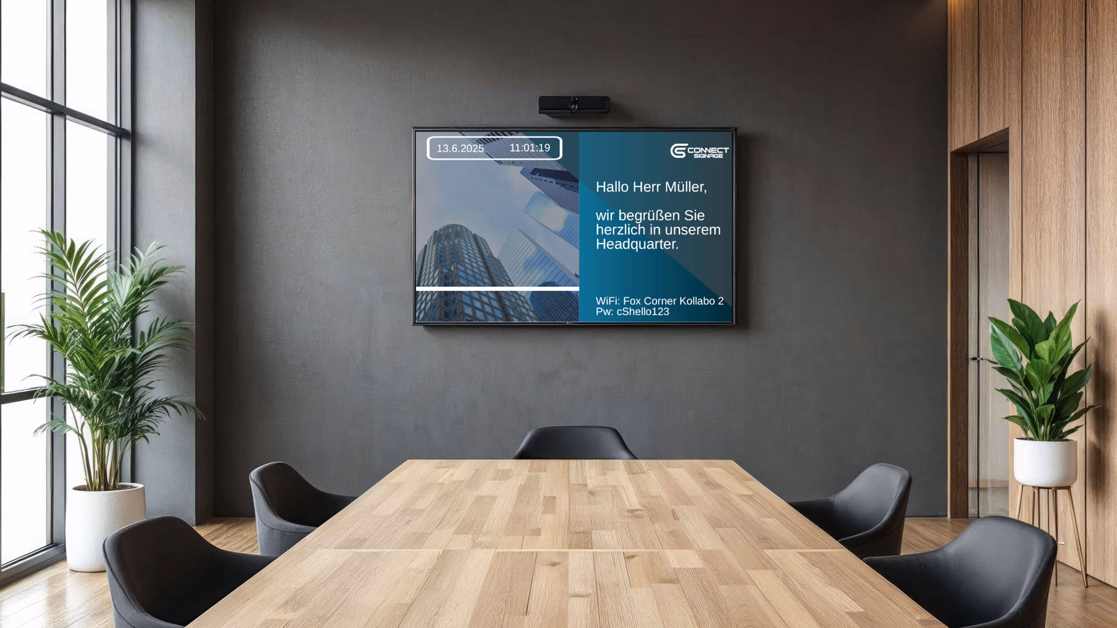 Moderner Besprechungsraum mit einem großen Holztisch, fünf dunklen Stühlen, zwei Zimmerpflanzen und einem Bildschirm an der grauen Wand.