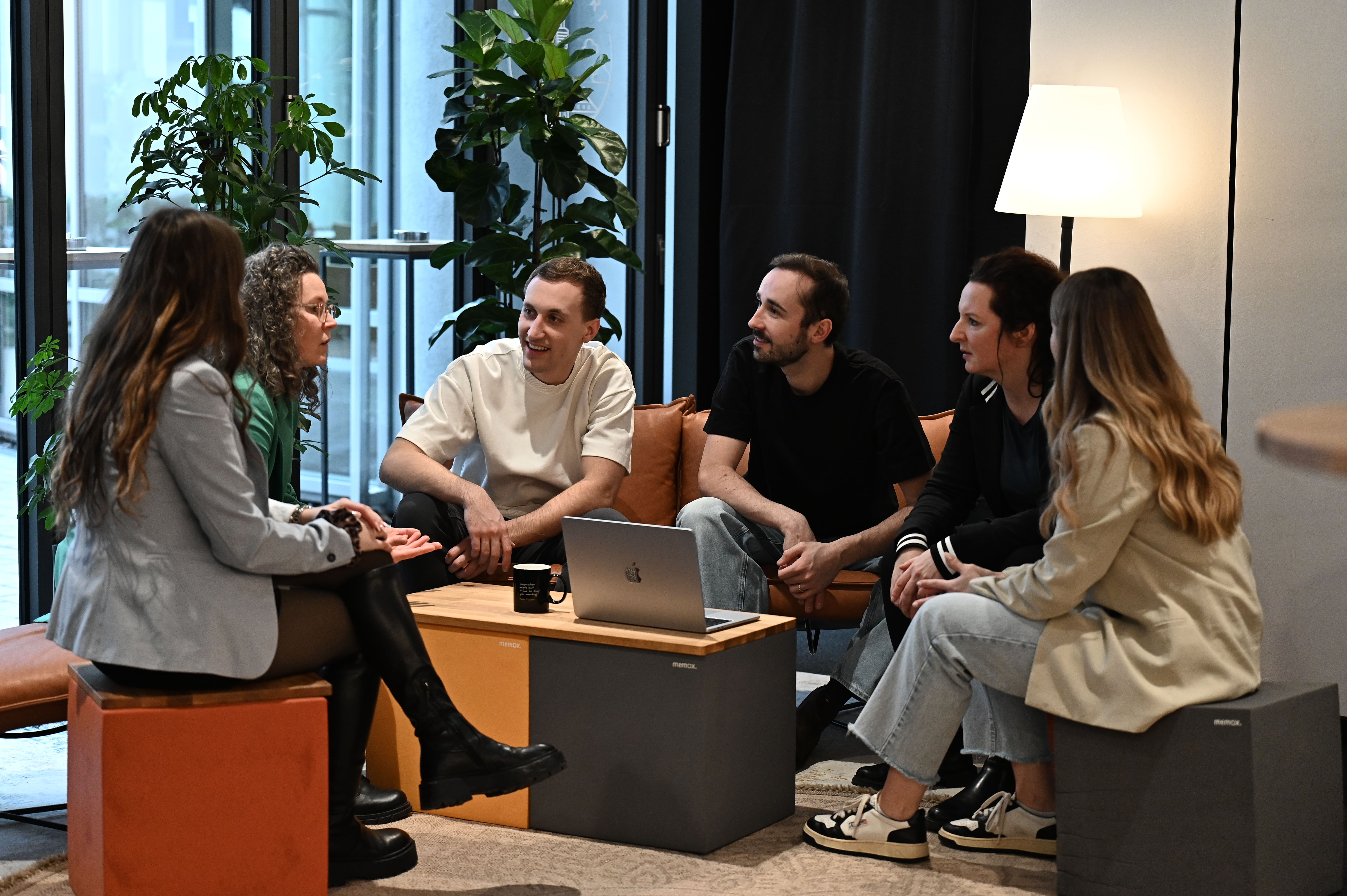 Sechs junge Erwachsene sitzen in einem modernen Bürobereich um einen Couchtisch mit Laptop und Tasse und führen ein Gespräch.