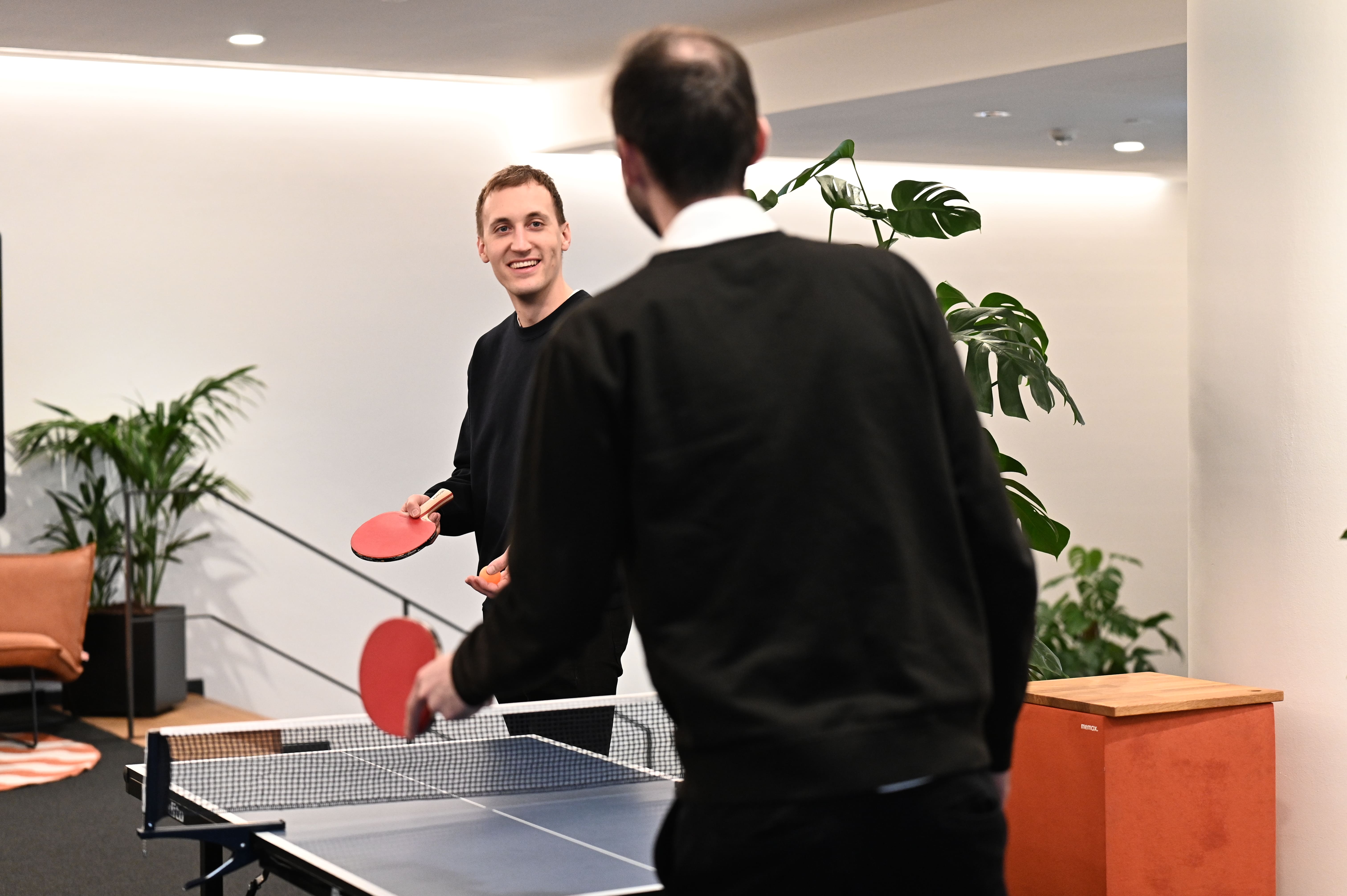 Zwei Männer spielen Tischtennis in einem modernen Innenraum mit Pflanzen und Sitzbereich.