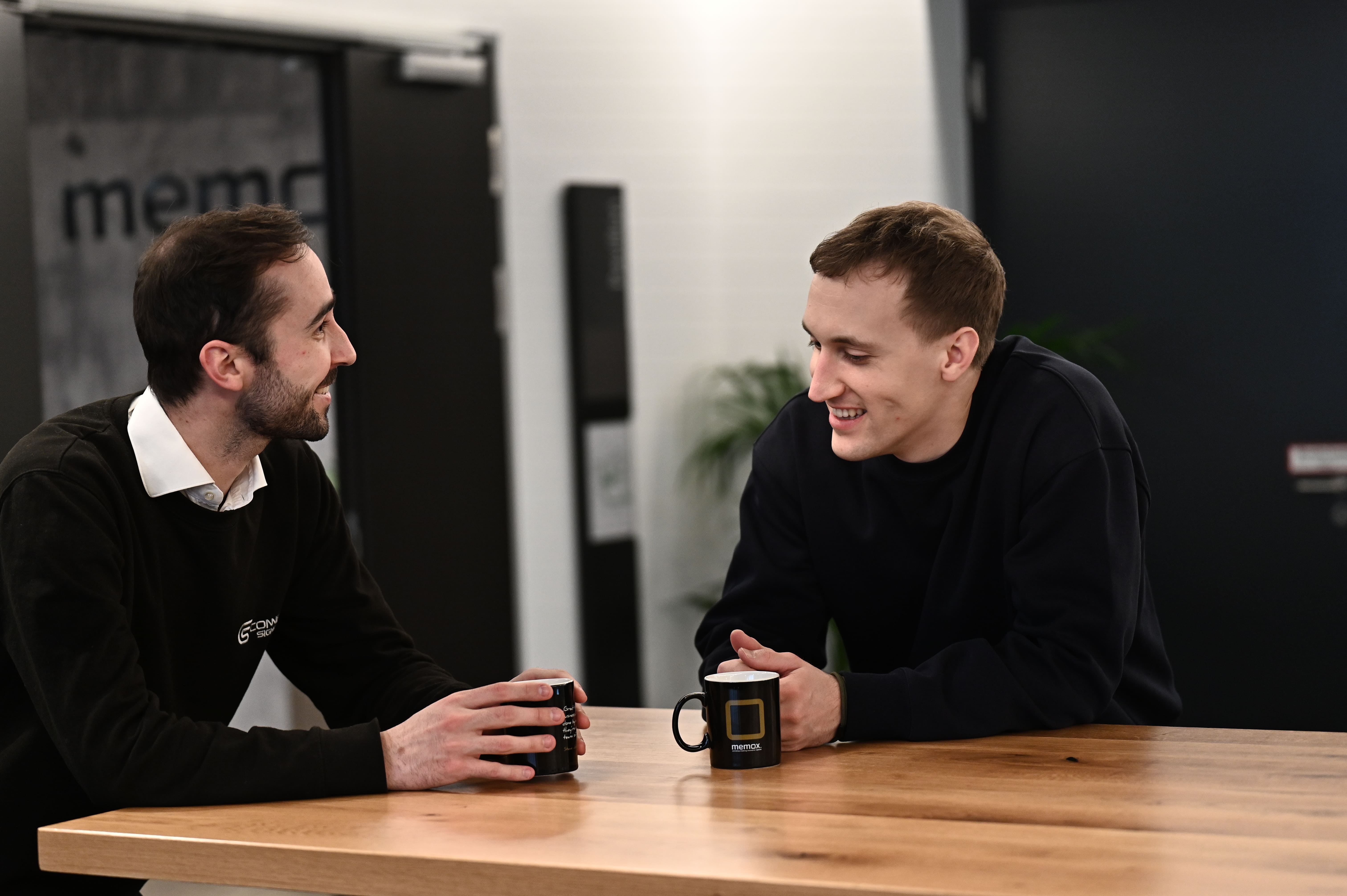 Zwei Männer unterhalten sich und lächeln bei einer Kaffeepause an einem Holztisch.