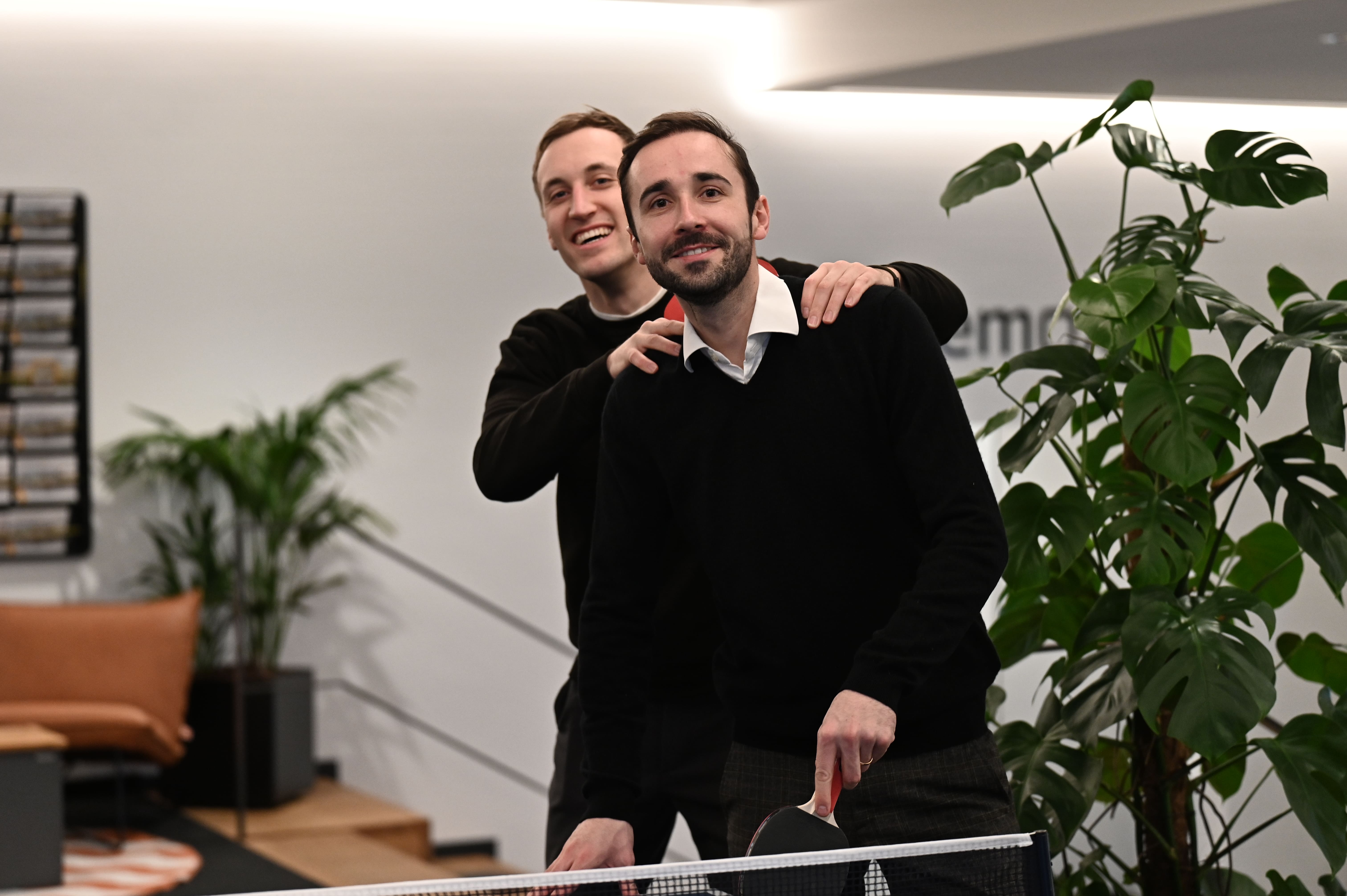 Zwei Männer lachen zusammen beim Tischtennis spielen in einem modernen Raum mit Pflanzen.
