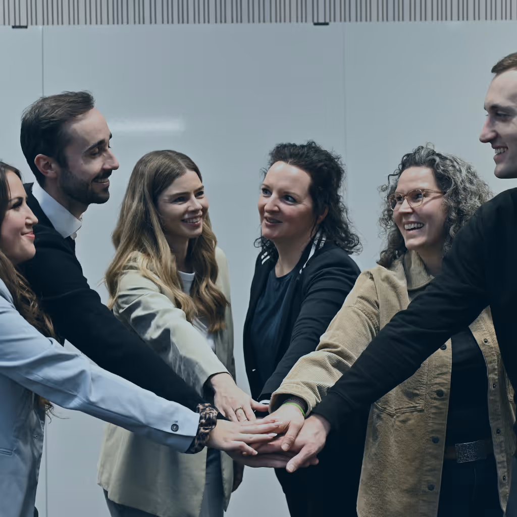 Sechs lächelnde Menschen legen ihre Hände zusammen als Zeichen von Teamarbeit.