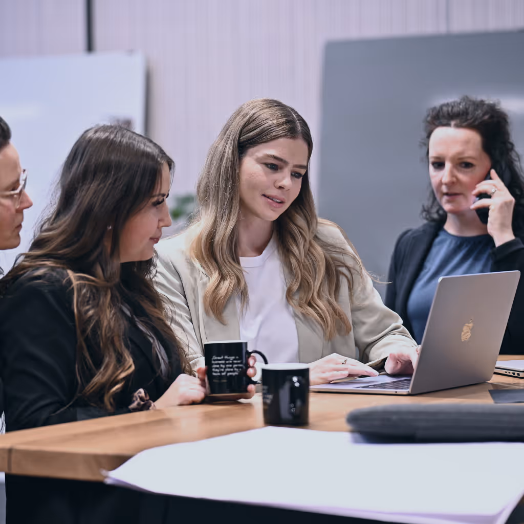 Vier Frauen bei einer Besprechung an einem Tisch, eine benutzt einen Laptop, eine andere telefoniert daneben.