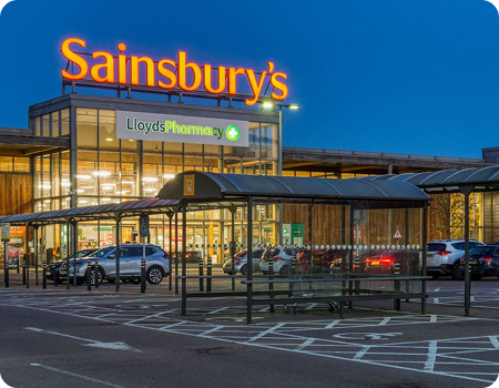Sainsbury's supermarket exterior at dusk with parked cars and a covered shopping cart area.