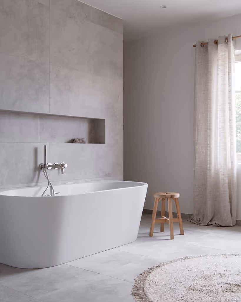 Minimalist bathroom with a white freestanding bathtub, wall-mounted faucet, wooden stool, beige curtains, and a round textured rug.