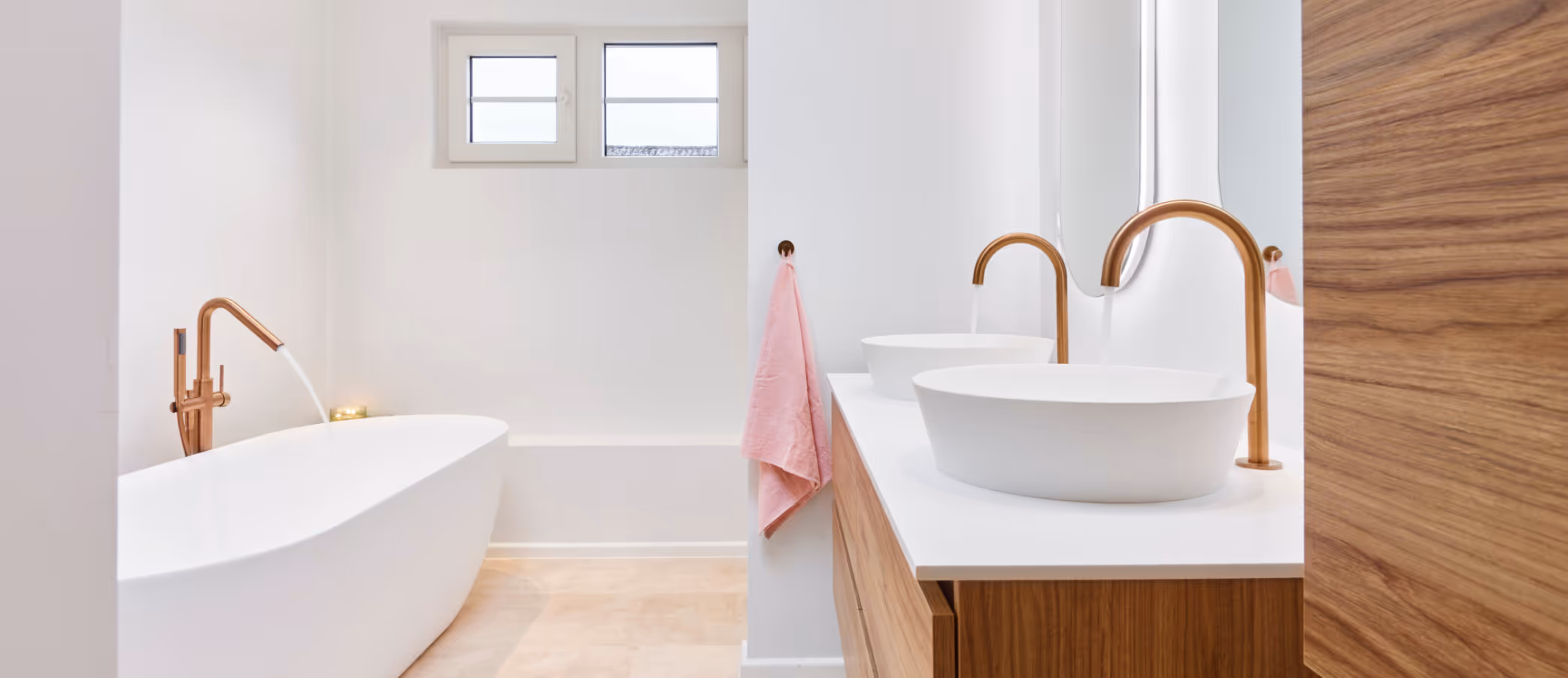 Moderne badkamer met een witte vrijstaande badkuip, dubbele wastafels met ronde witte kommen en koperkleurige kranen, houten kast en een roze handdoek aan de muur.