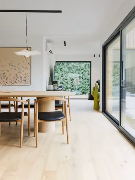 Moderne eetkamer met licht houten vloer, houten tafel met zwarte stoelkussens, grote ramen en uitzicht op groene tuin.