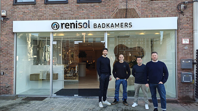 Vier mannen poseren voor de gevel van renisol badkamers winkel met glazen deuren en een bakstenen muur.
