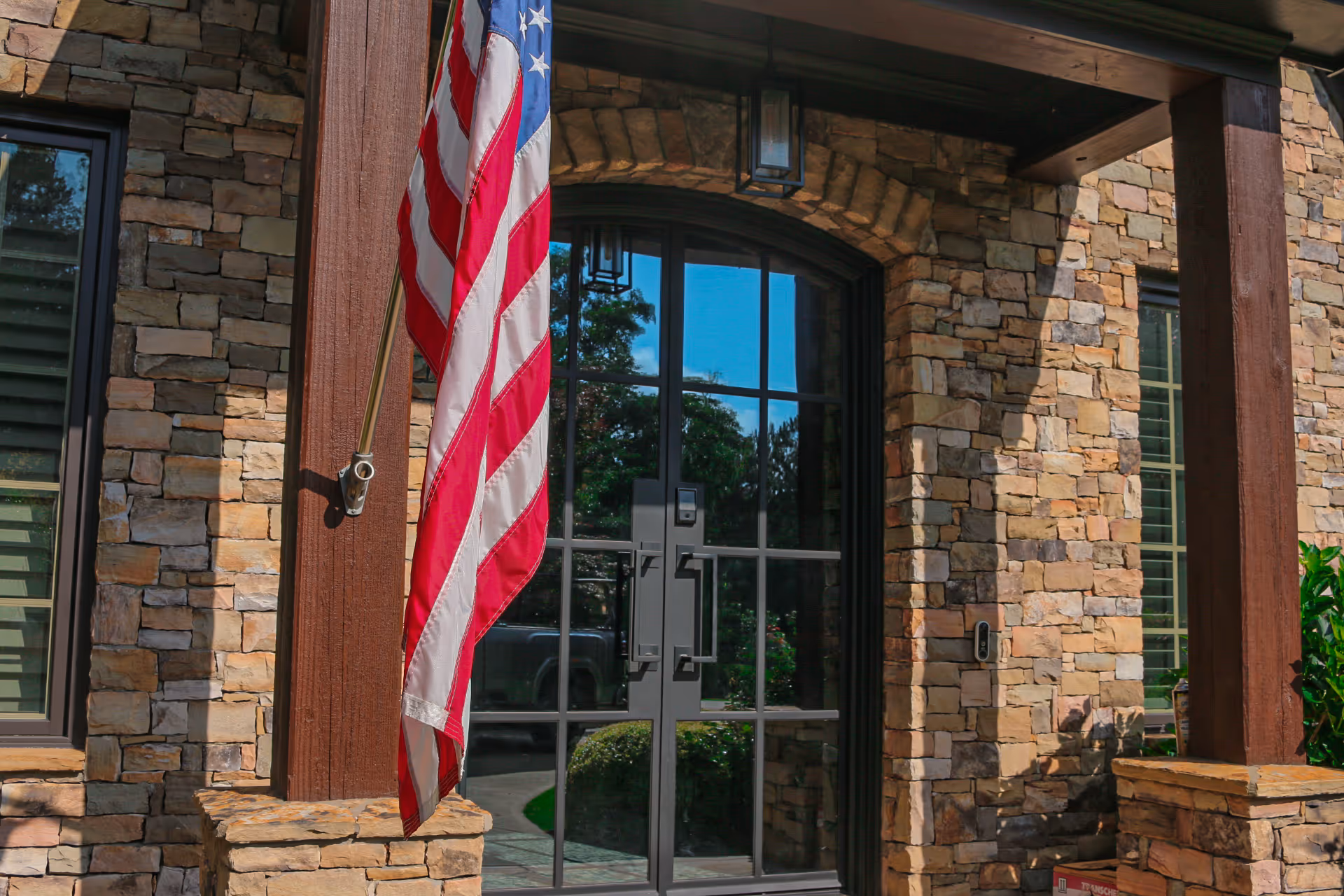 Stone house entrance with a glass double door reflecting trees and an American flag hanging on a wooden post.