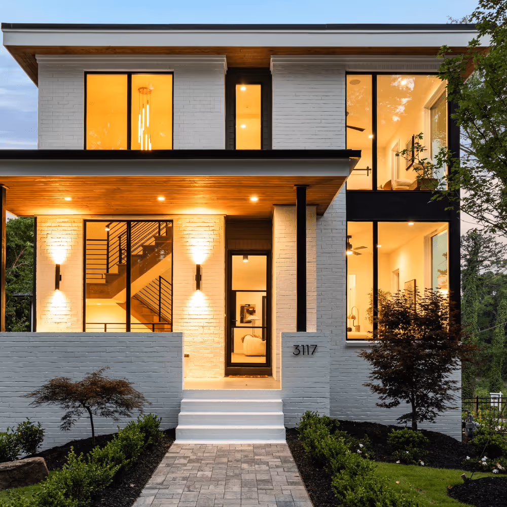 Modern two-story white brick house with large windows, warm interior lighting, and landscaped front yard.