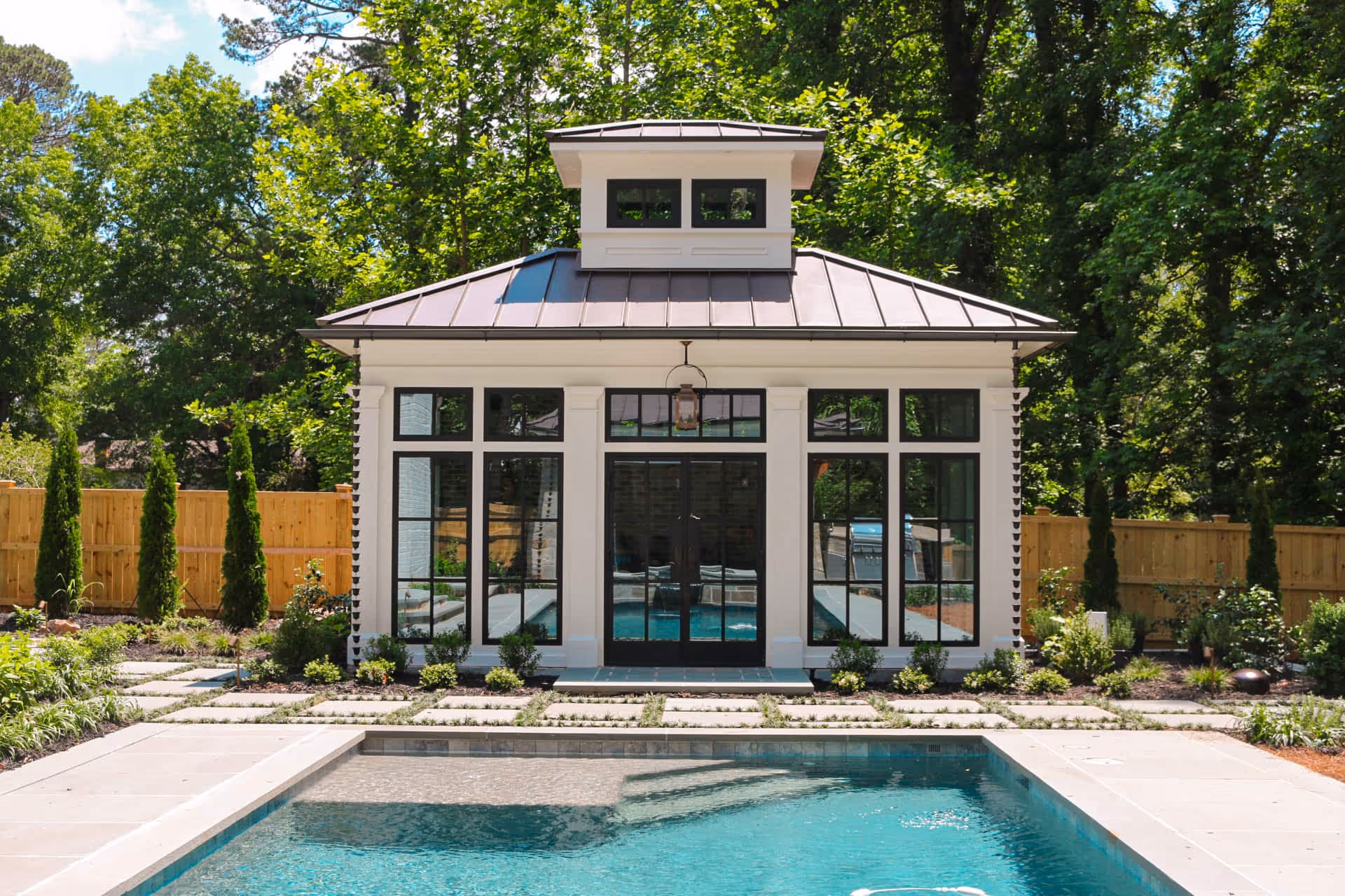 Modern white pool house with large black-framed windows and a small tower roof, situated behind a clear blue swimming pool and surrounded by greenery and a wooden fence.