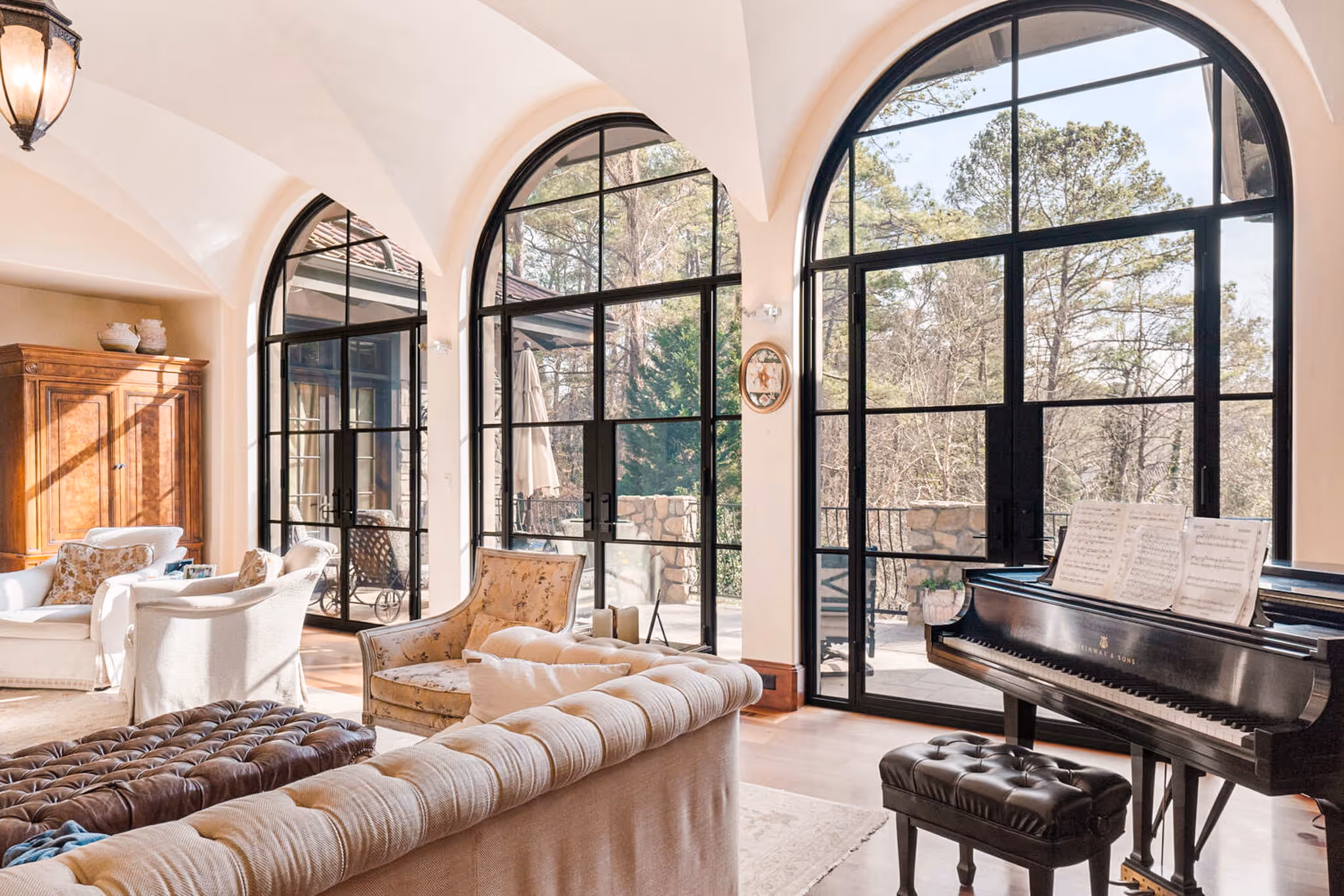 Bright living room with arched floor-to-ceiling windows, beige sofas, floral armchair, and a black grand piano with sheet music.