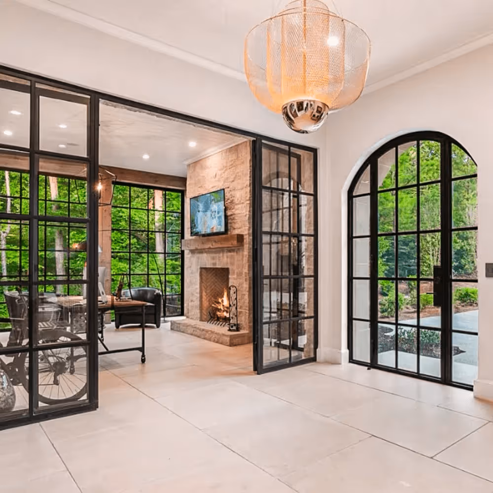 Modern living area with large black framed glass doors, a stone fireplace with fire lit, and a view of green trees outside.