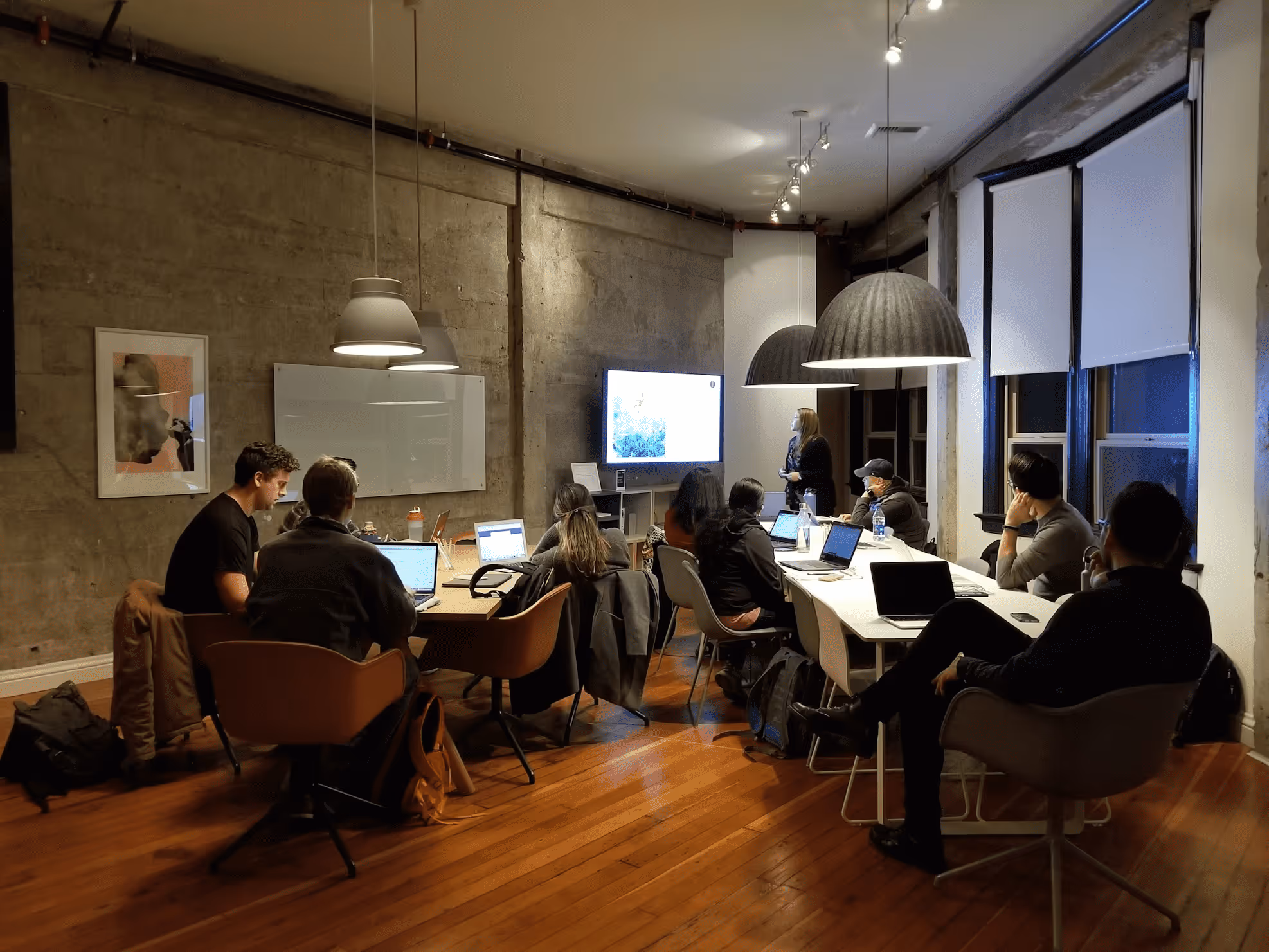 Group of people sitting around two tables in a modern office with laptops, watching a presentation on a wall-mounted screen.