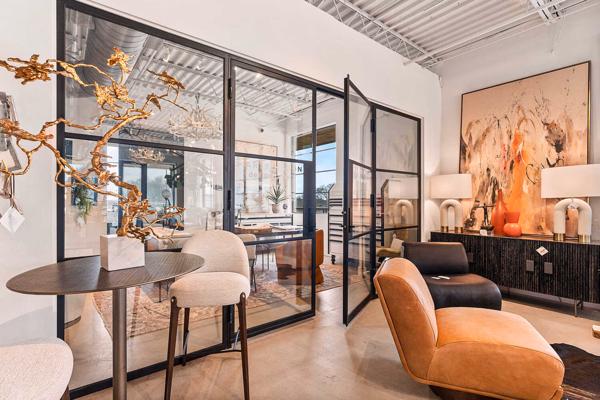 Modern living room with two tan and black leather chairs, a wooden table with a gold decorative sculpture, glass doors, and abstract painting on the wall.