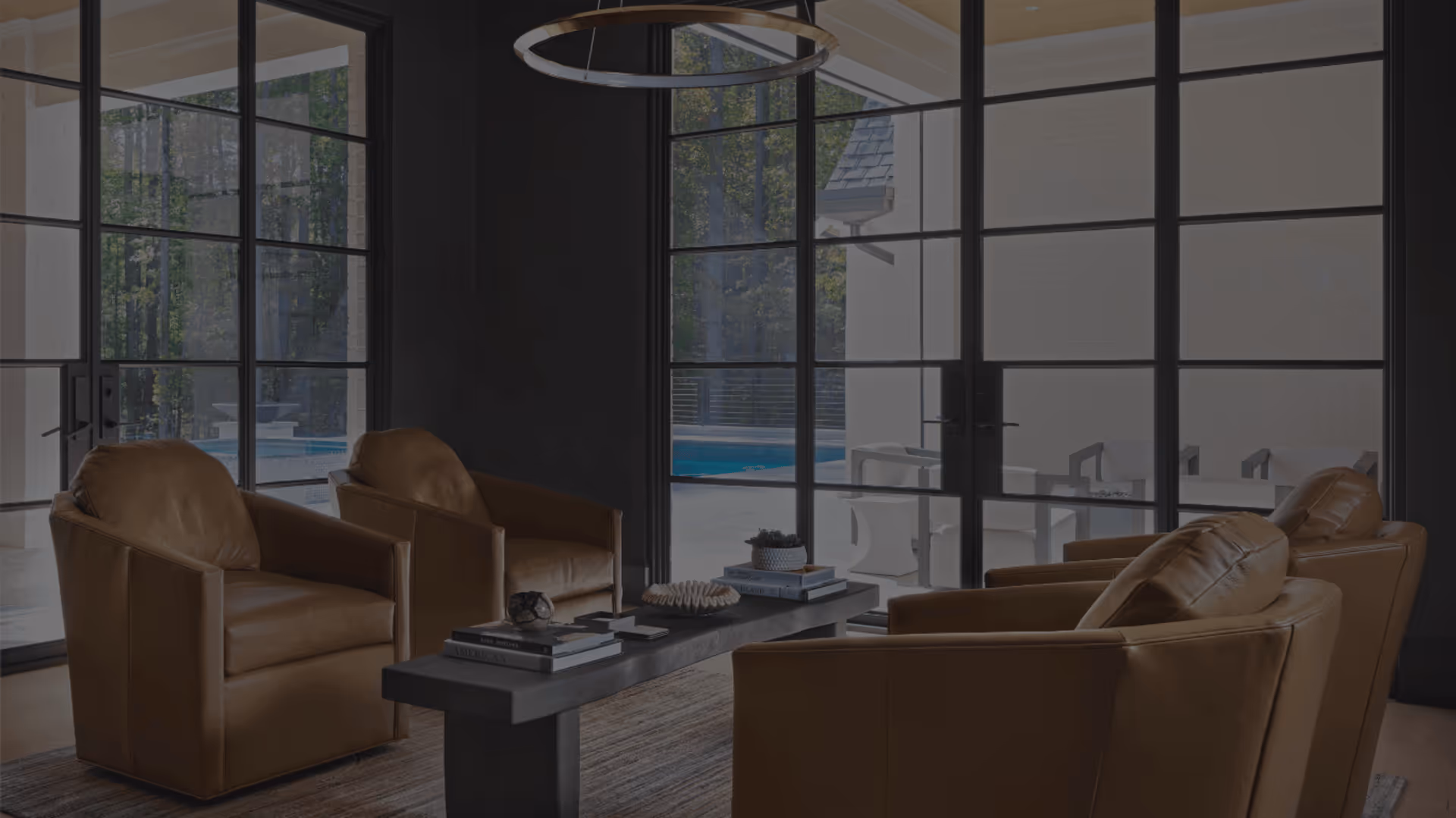 Modern living room with four brown leather armchairs around a black rectangular coffee table, large black-framed windows showing a pool and patio outside.