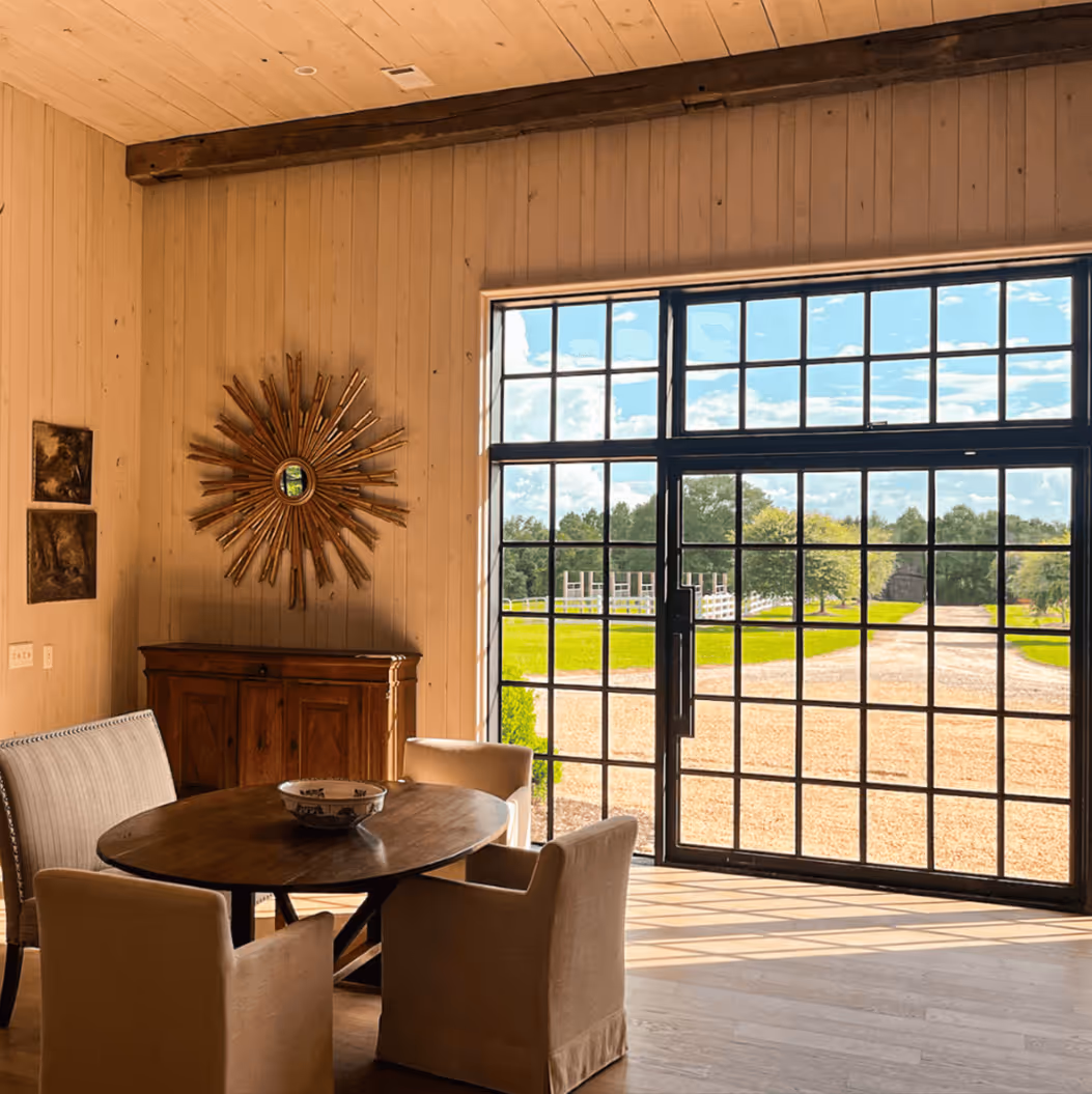 Dining area with round wooden table, four beige chairs, sunburst mirror on wood-paneled wall, and large window showing green outdoor landscape.