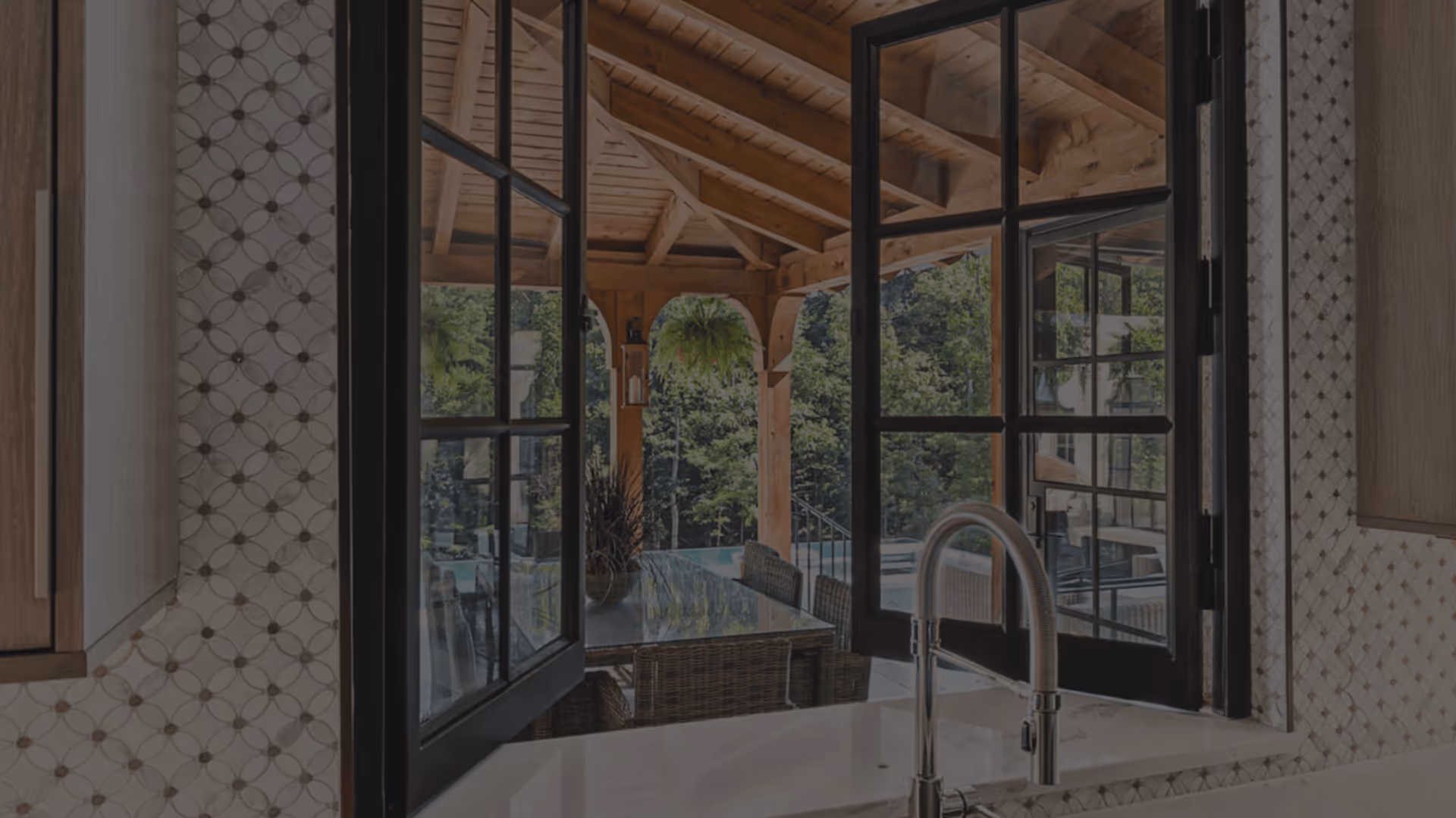 View through open black framed windows from kitchen sink to an outdoor covered patio with wicker chairs, glass table, hanging fern, and lush green trees in background.