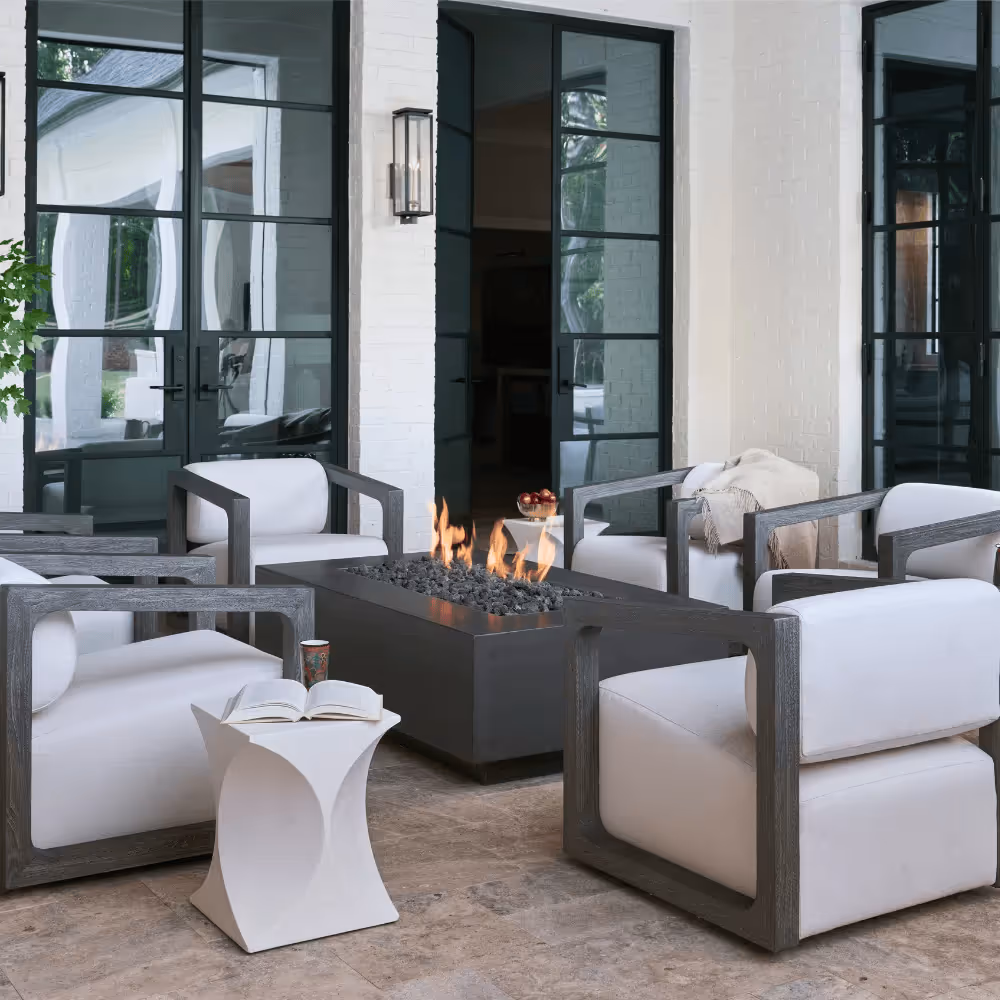 Outdoor seating area with four modern armchairs around a rectangular fire pit, a small white side table with an open book and cup, and large glass doors in the background.