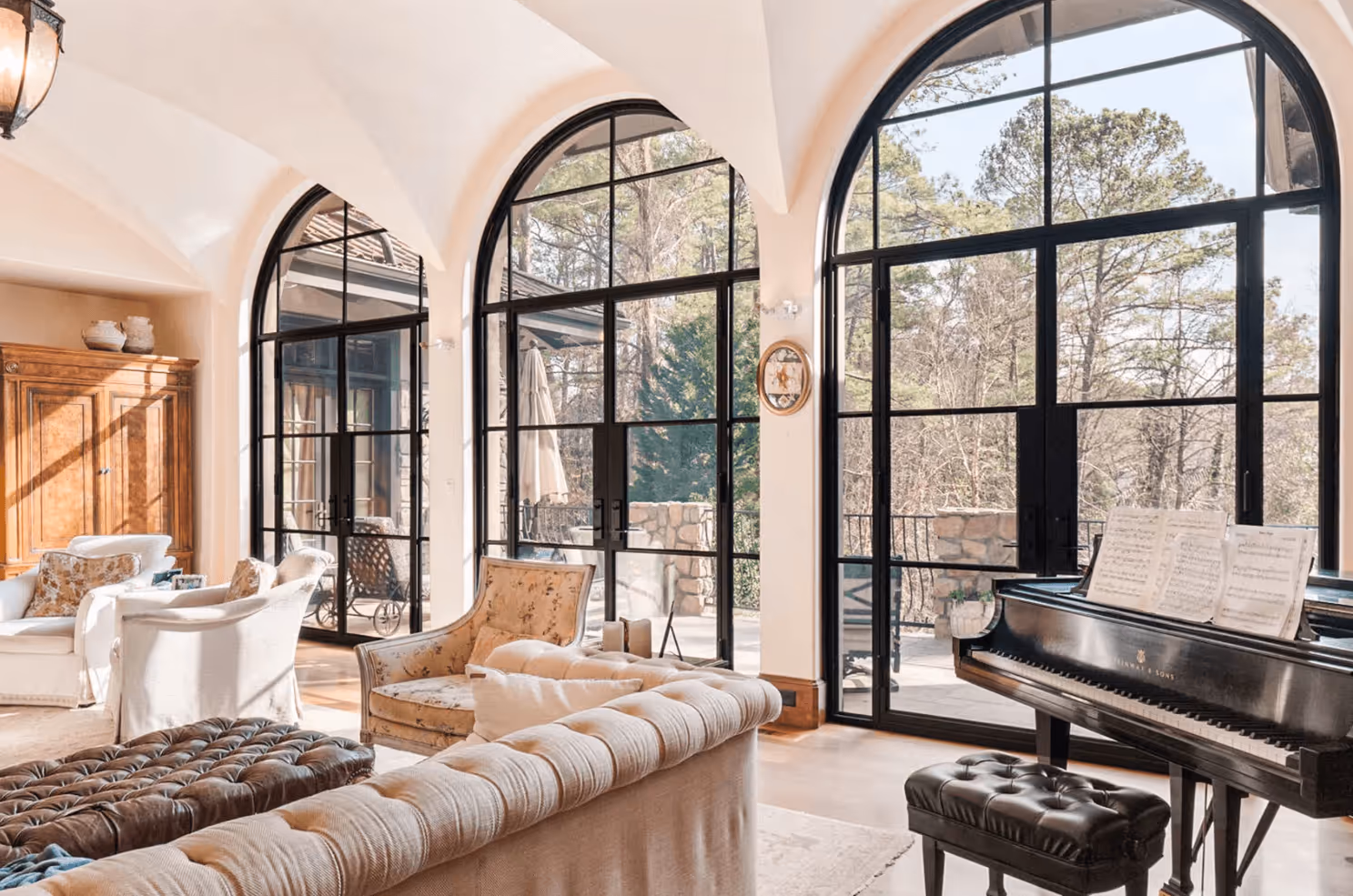Bright living room with large arched windows, beige sofas, armchairs, and a black grand piano with sheet music.