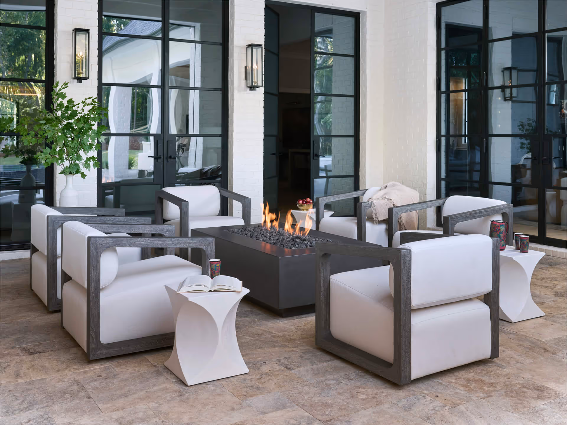 Modern outdoor seating area with six white cushioned armchairs around a rectangular fire pit on a tiled floor.