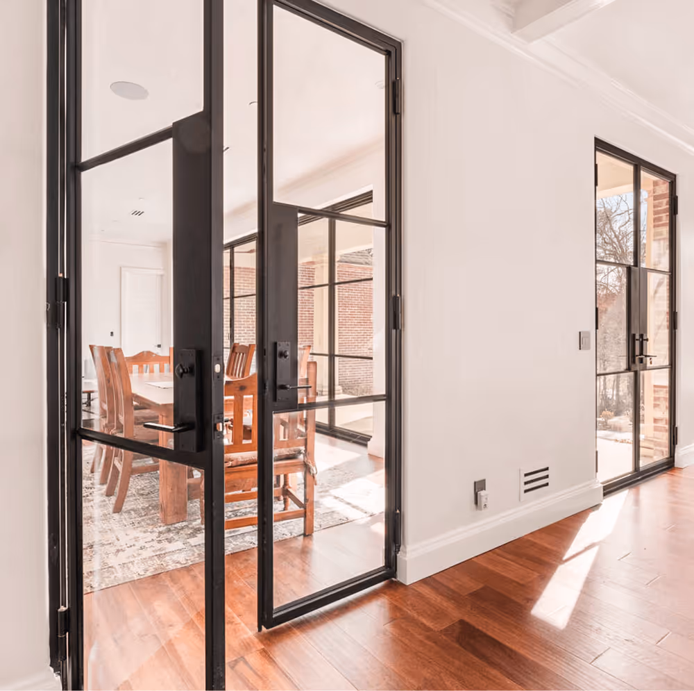 Black-framed glass double doors opening to a dining area with wooden chairs and a patterned rug, sunlight shining on hardwood floor.