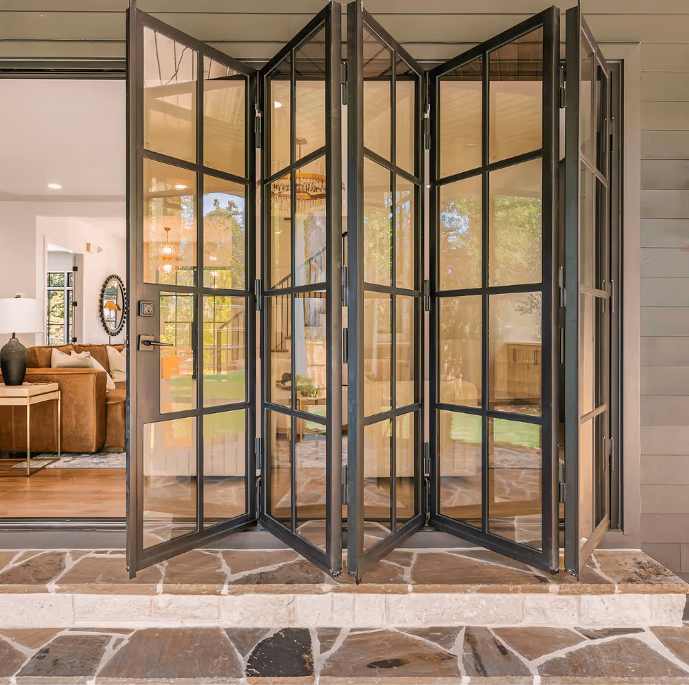 Open black metal framed folding glass doors leading to a stone patio with a view of a cozy living room inside.