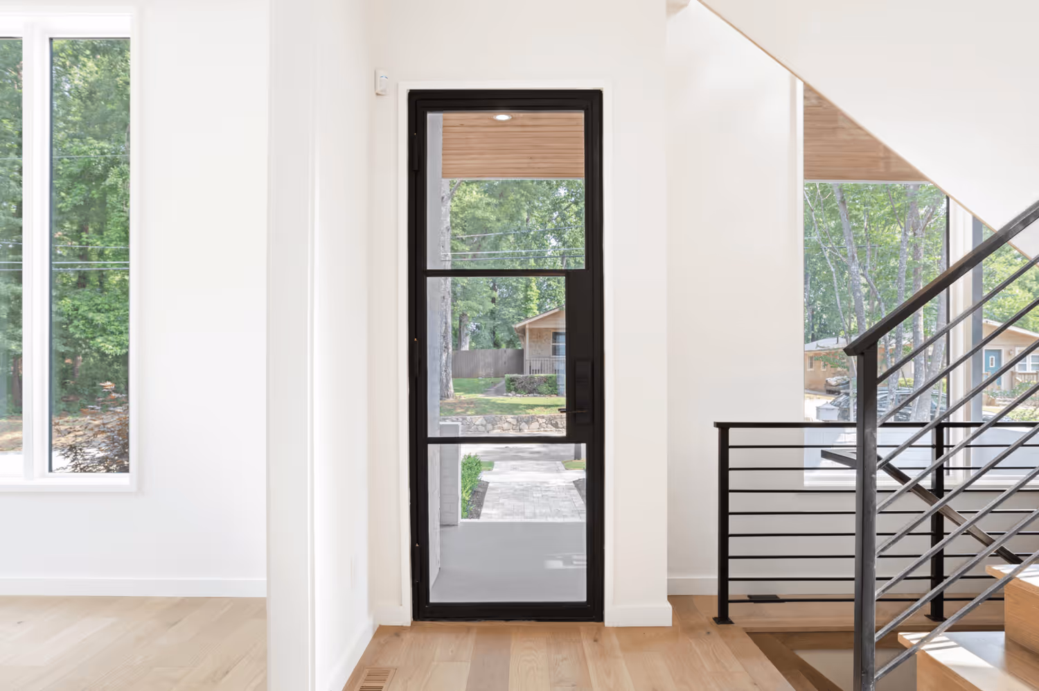 Modern interior with a black-framed glass door showing a view of a driveway and trees outside, next to a staircase with black railings.