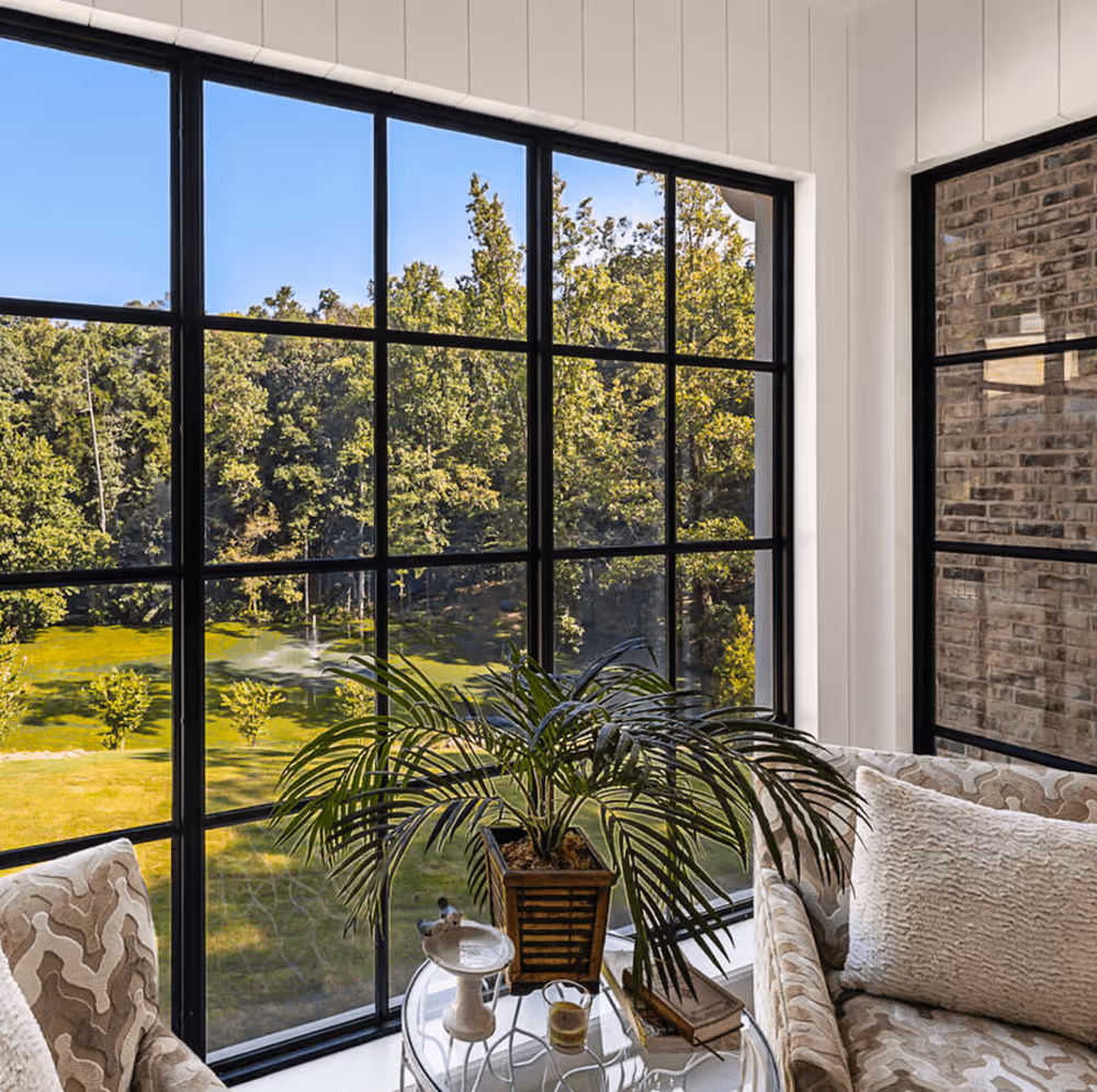 Living room corner with large black-framed windows overlooking a green yard with trees, featuring patterned armchairs, a glass-top table with a potted plant, books, and a candle.