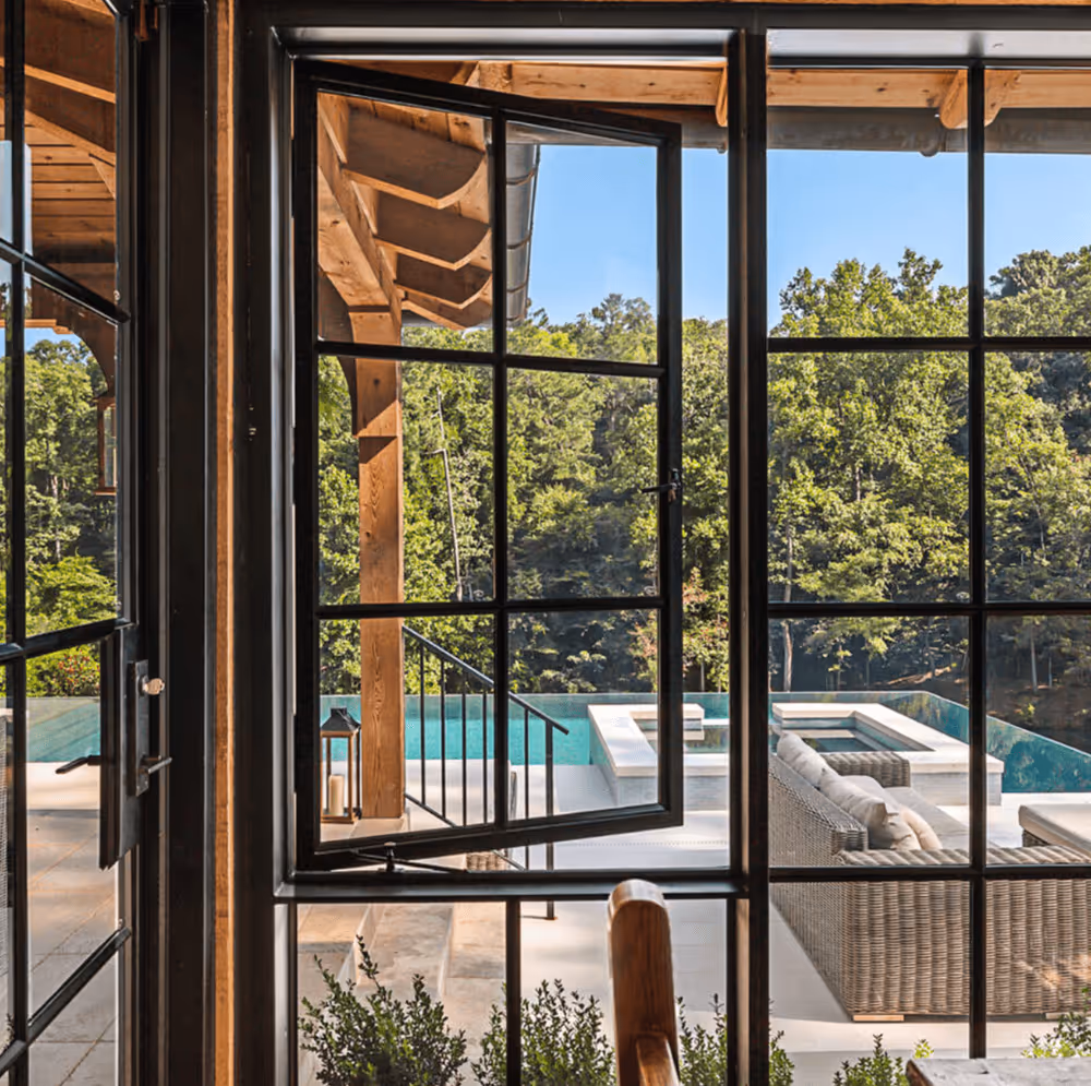 View through black-framed glass doors of a patio with wicker seating and a pool surrounded by trees under a clear blue sky.