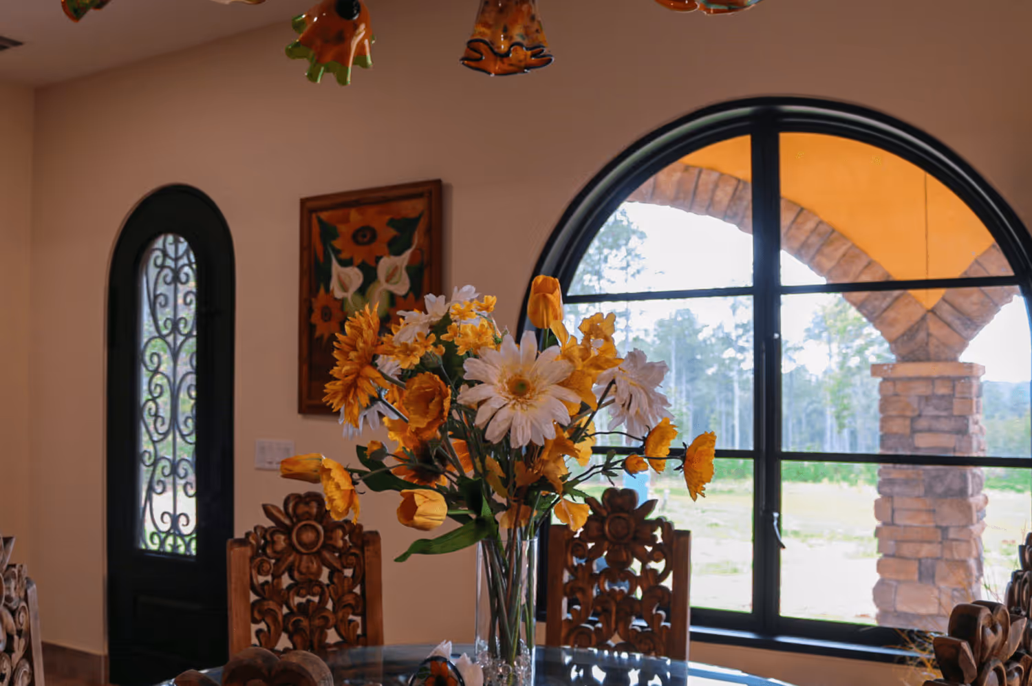 Vase with yellow and white flowers on a glass table in a dining area with carved wooden chairs, an arched window, and a decorative door.