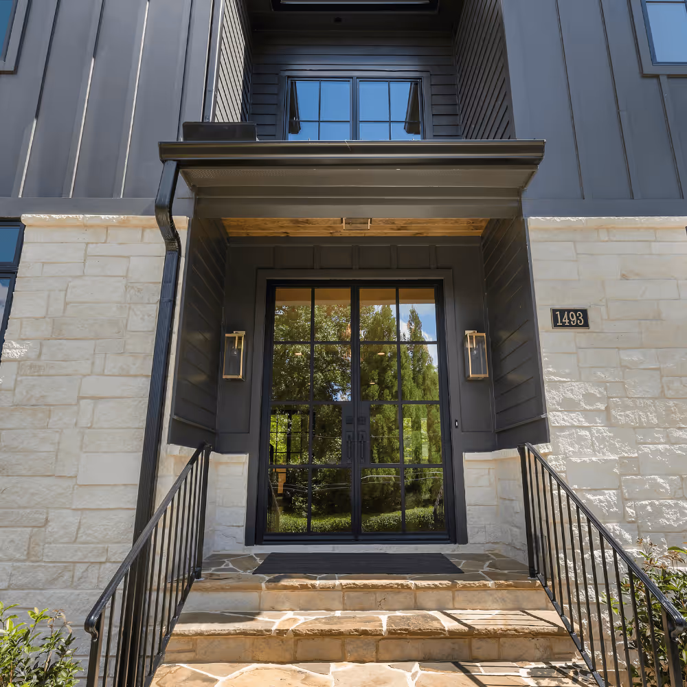 Modern black steel double door with glass panes and sidelight lanterns on stone and dark siding exterior front entrance with steps and black railings.