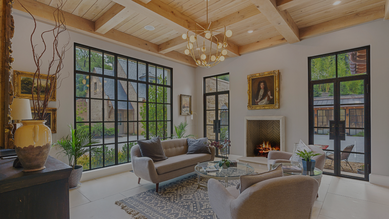 Cozy modern living room with large black-framed windows, gray sofas, a glass coffee table, a lit fireplace, and wooden ceiling beams.