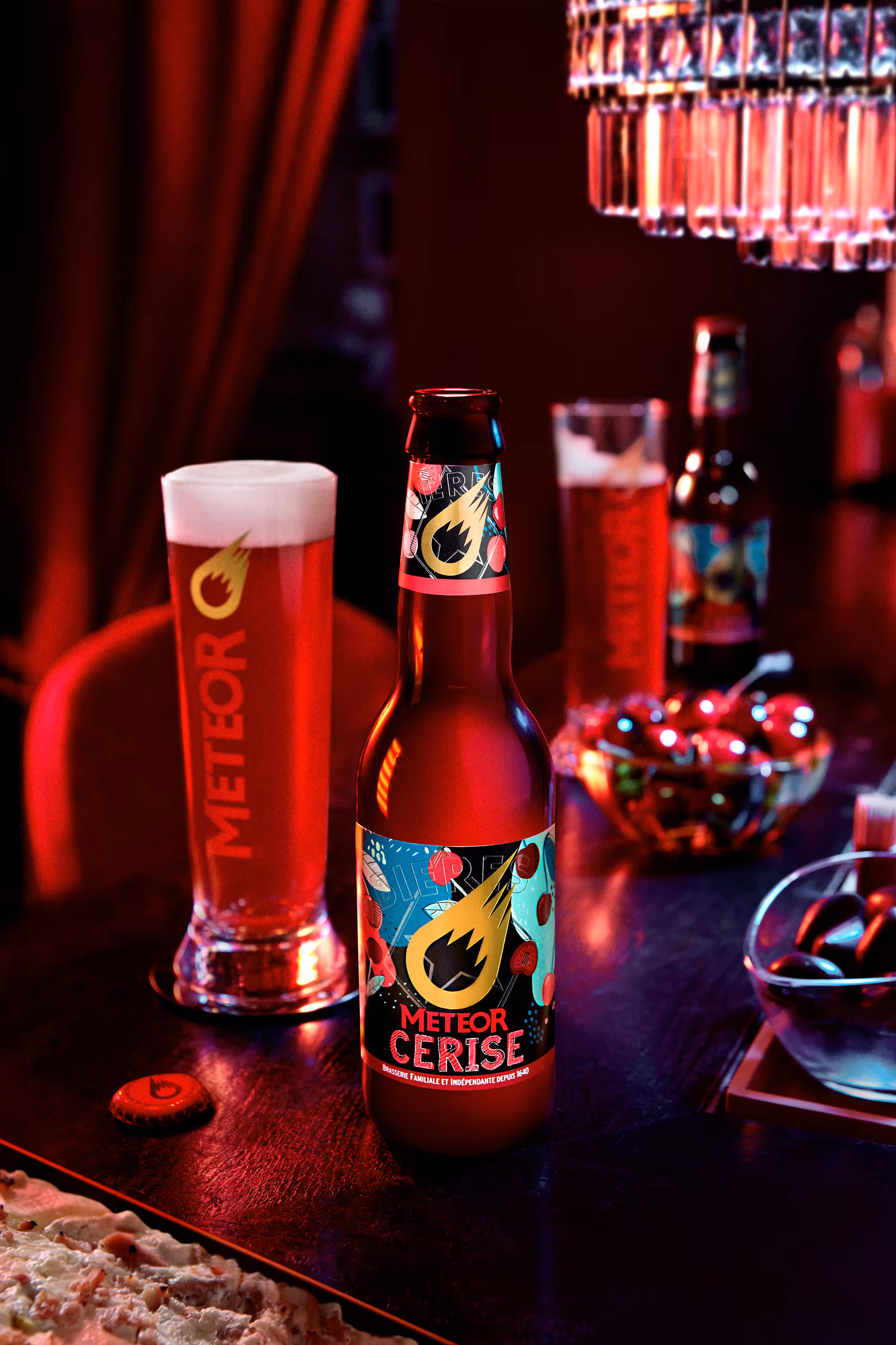 Bottle of Meteor Cerise cherry beer with a filled Meteor glass and a bowl of cherries on a dark table under warm lighting.