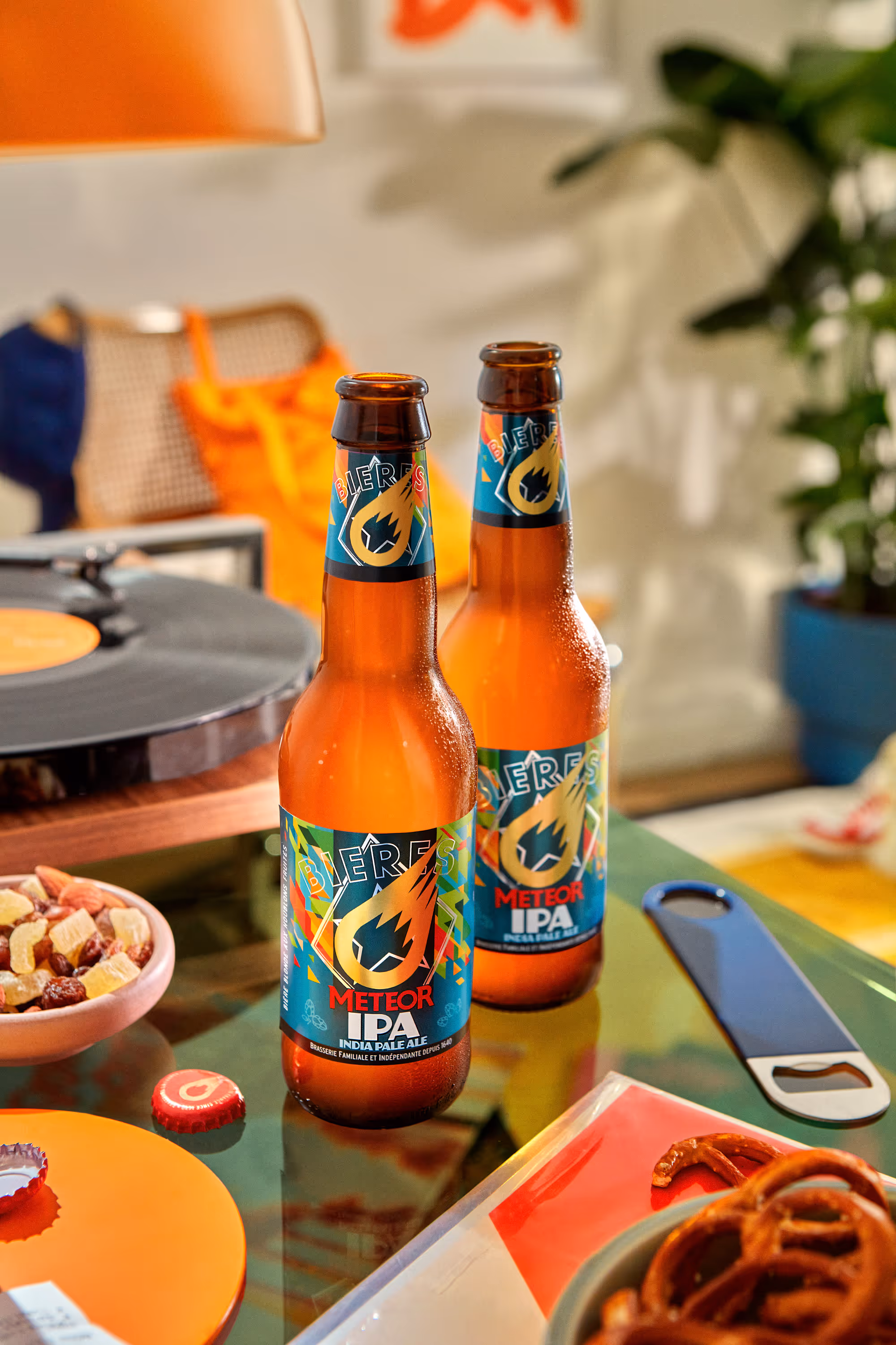Two bottles of Meteor IPA beer on a glass table surrounded by snacks, a bottle opener, and a vinyl record player.