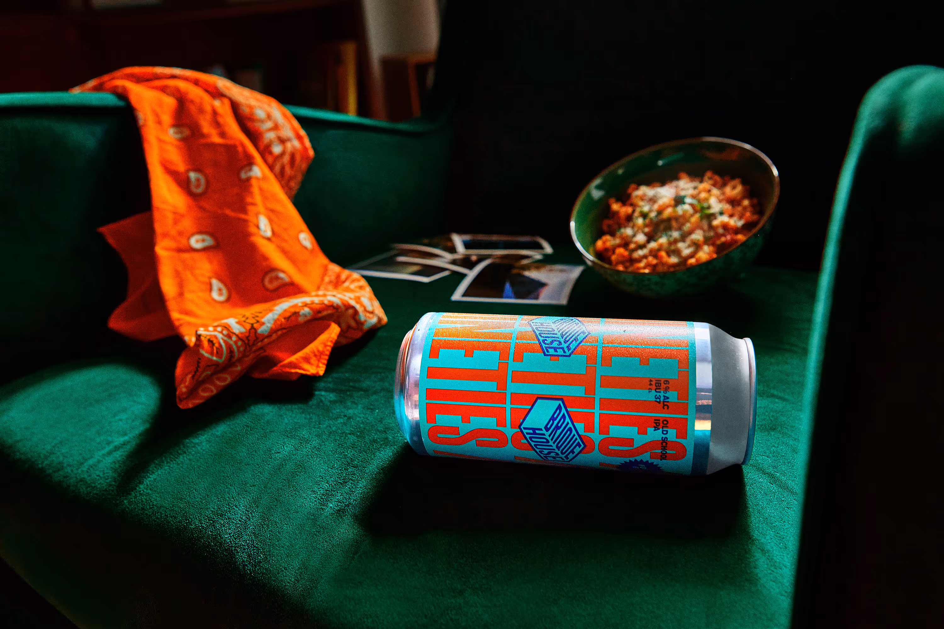 Orange bandana, a can of beer labeled 'Old School IPA', scattered photos, and a bowl of food on a green velvet couch.
