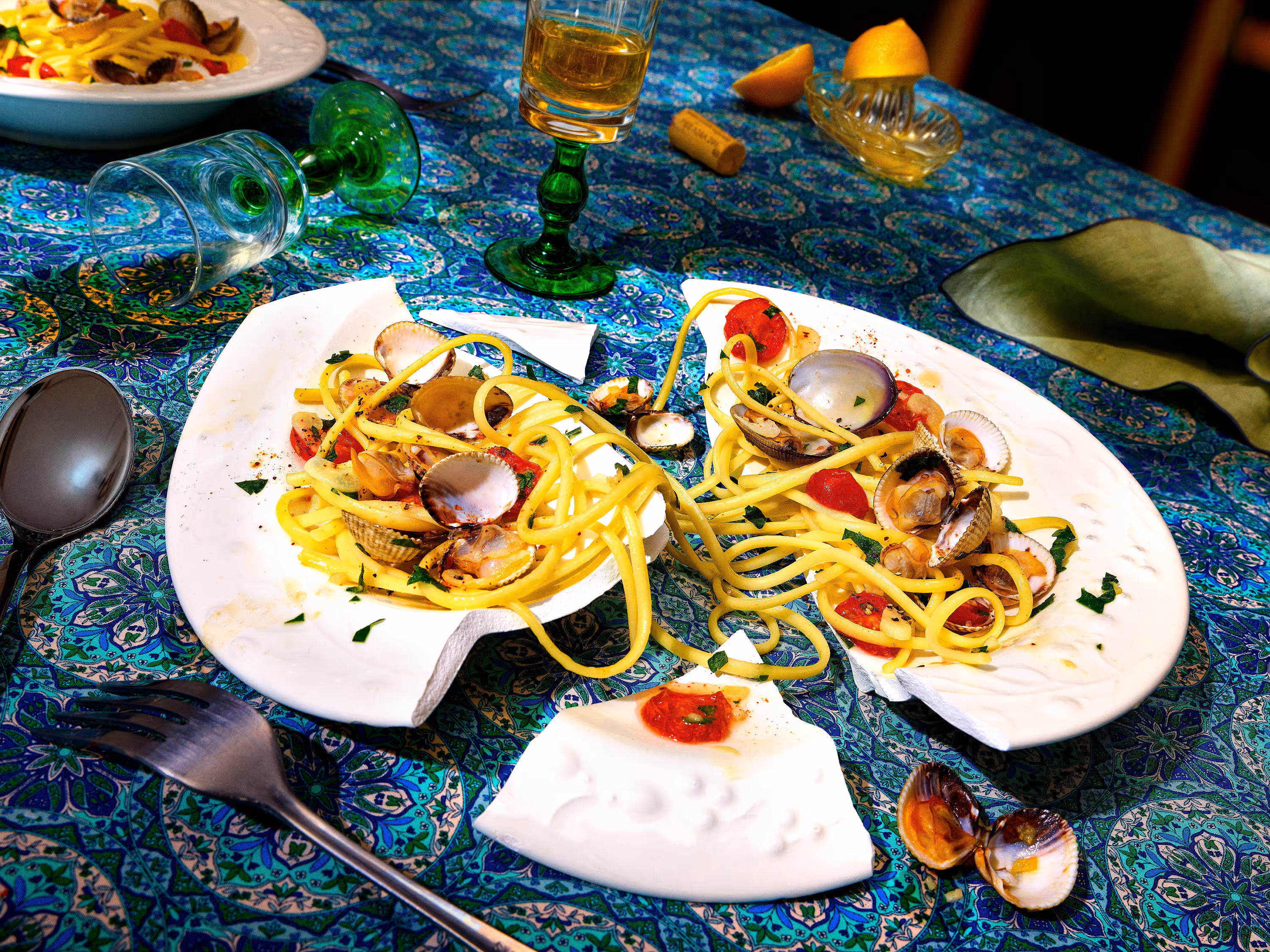 Broken white plate with spaghetti pasta, clams, and cherry tomatoes on a blue patterned tablecloth surrounded by green glassware, a fork, and lemon wedges.