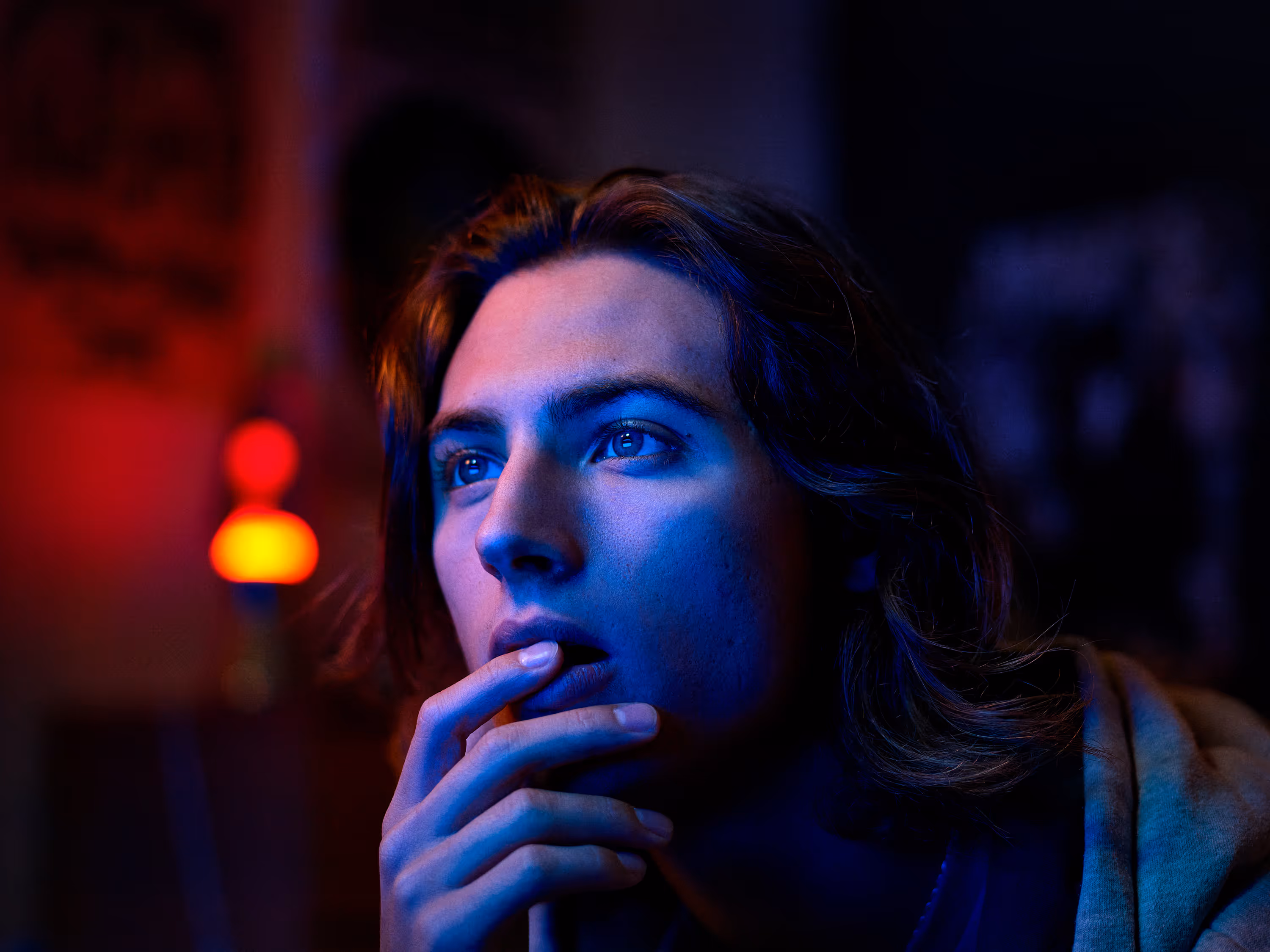 Young person with long hair illuminated by blue and red light, looking thoughtful with fingers near mouth.