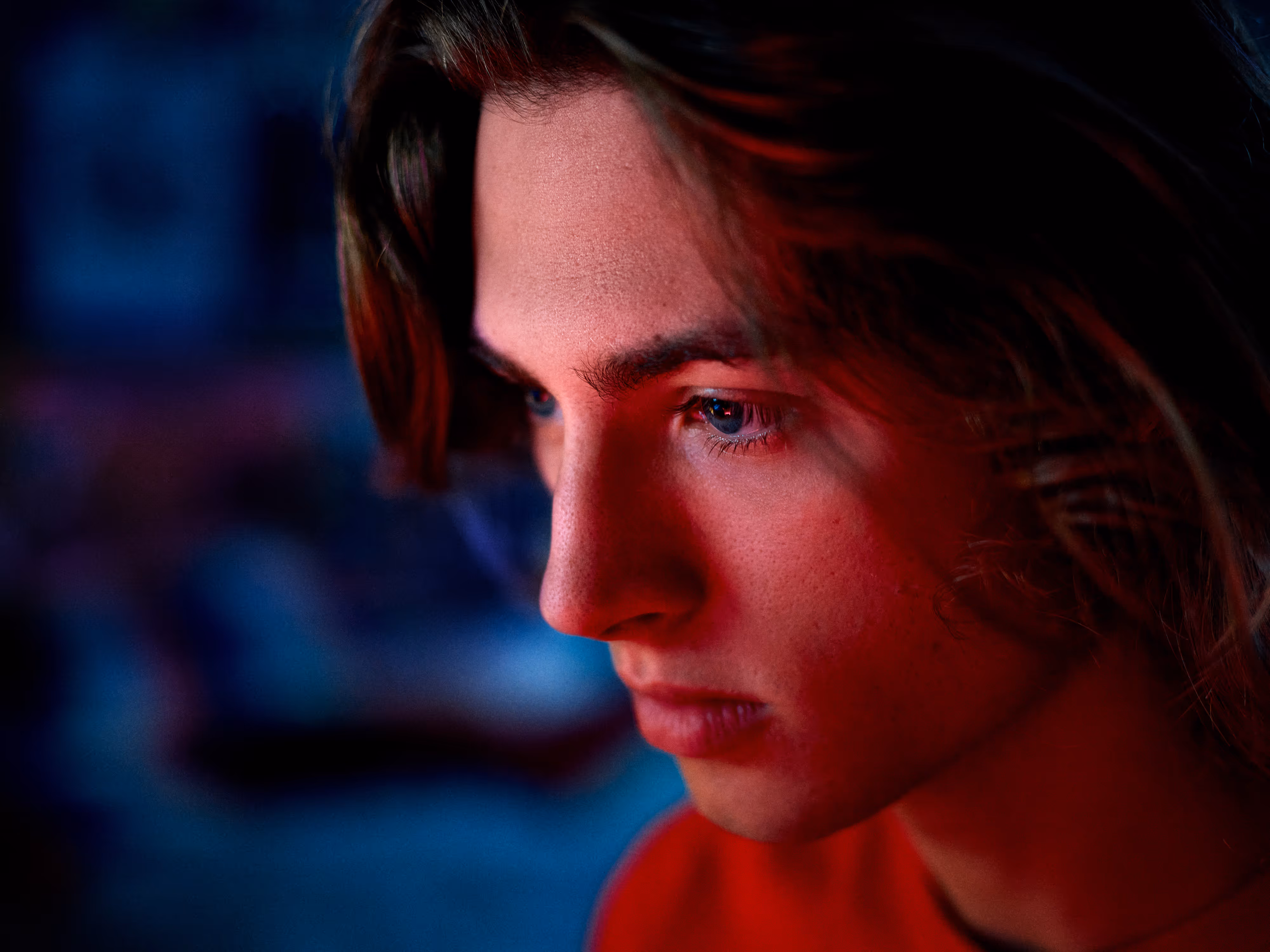 Close-up of a young person with long hair illuminated by red and blue lighting, looking thoughtfully to the side.