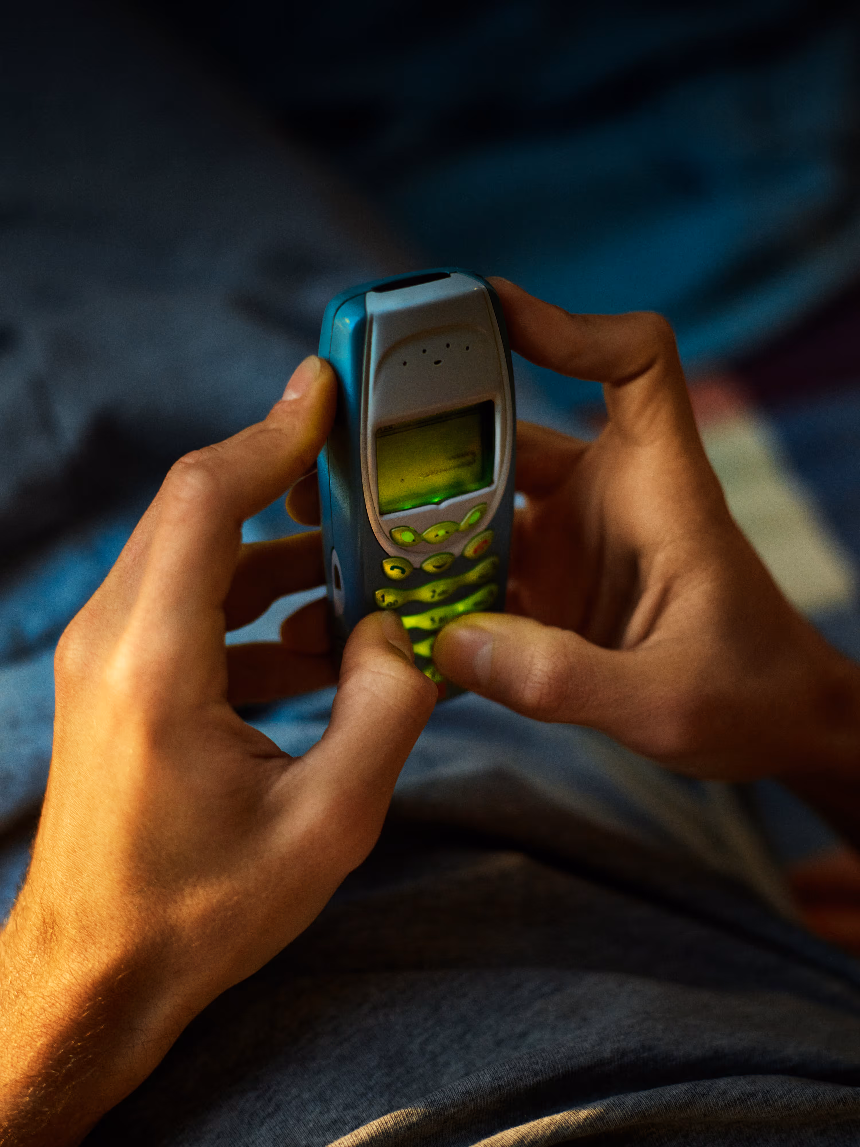 Hands holding and typing on an old blue mobile phone with a green backlit keypad.
