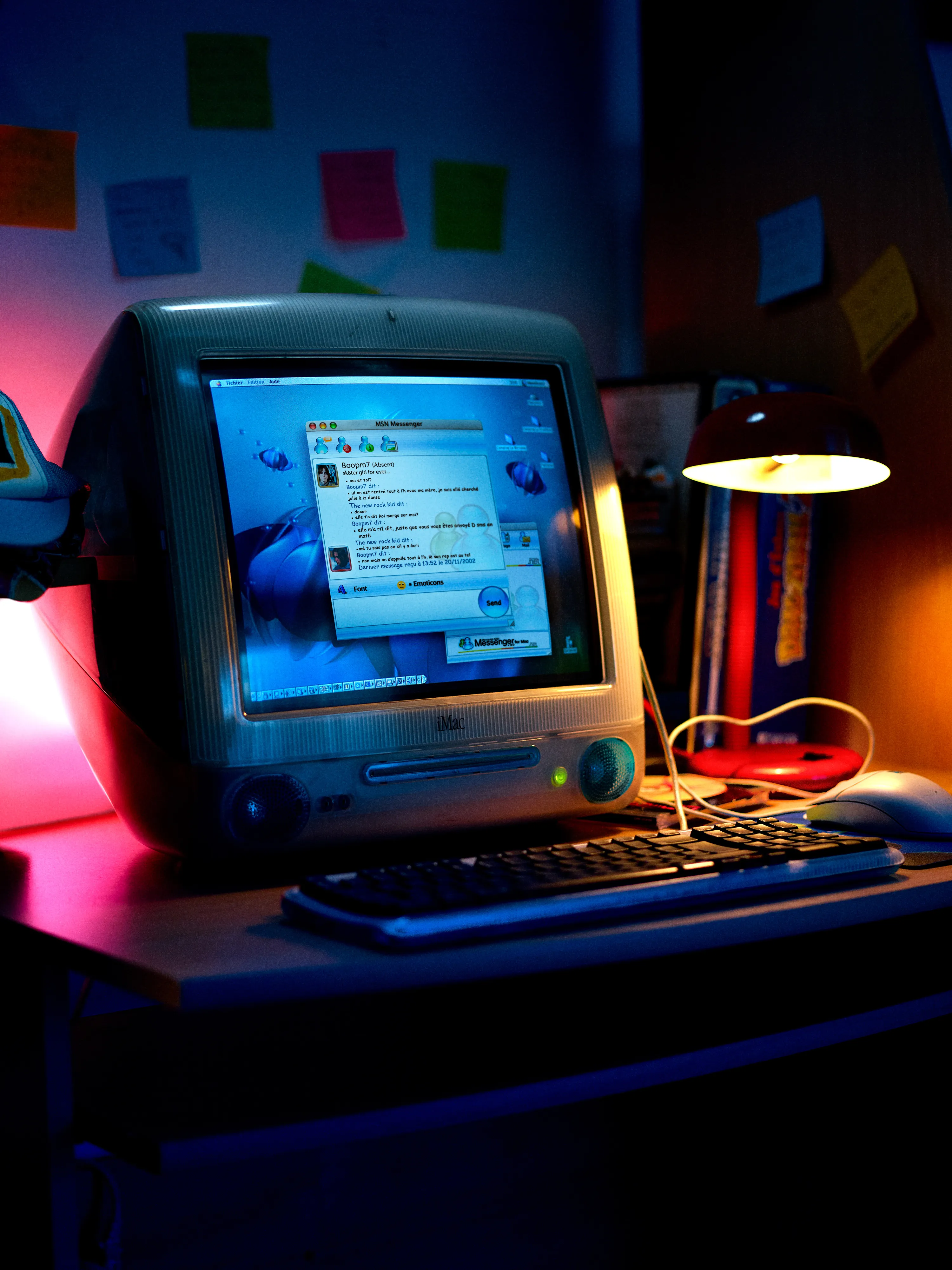 Vintage iMac computer on desk with MSN Messenger chat open, illuminated by a desk lamp and colorful sticky notes on the wall.